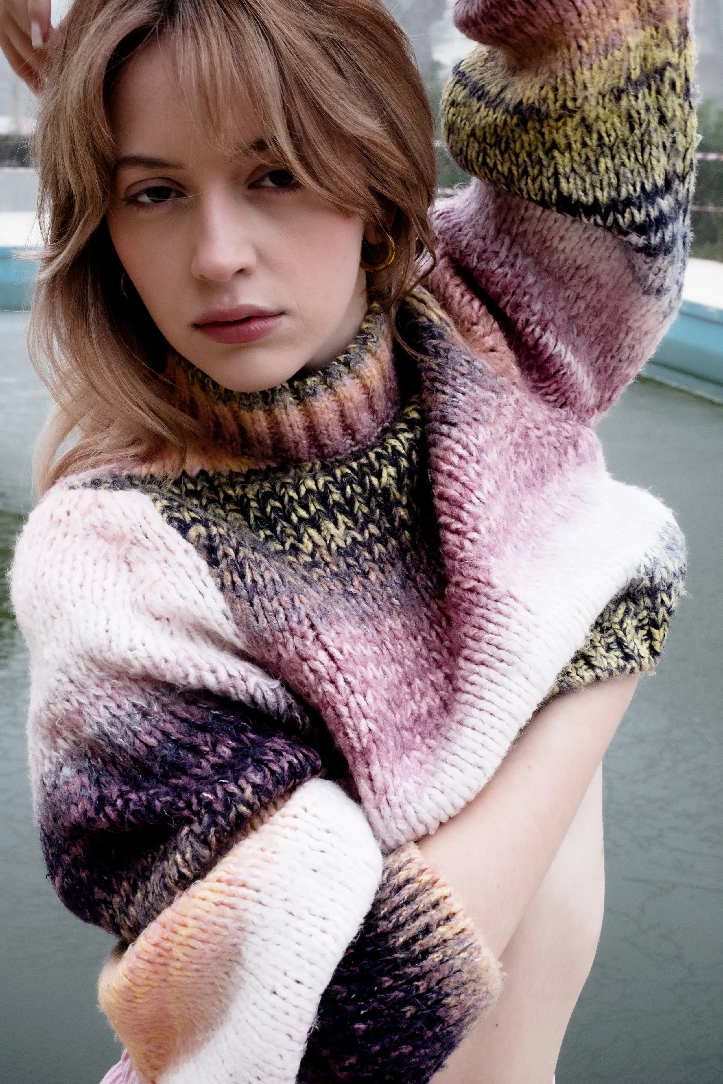 A woman with short, messy, reddish hair and gold hoop earrings is wearing a colorful, chunky knit sweater and is holding her arm across her chest in front of a body of water.