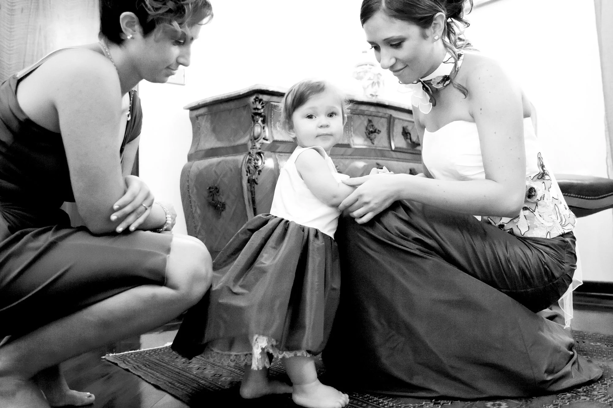 A young girl in a dress standing on a rug, with two women helping her, one on each side. The women are kneeling on the floor, holding the girl's dress, and they are dressed up, indicating a formal occasion, possibly a wedding or special event.