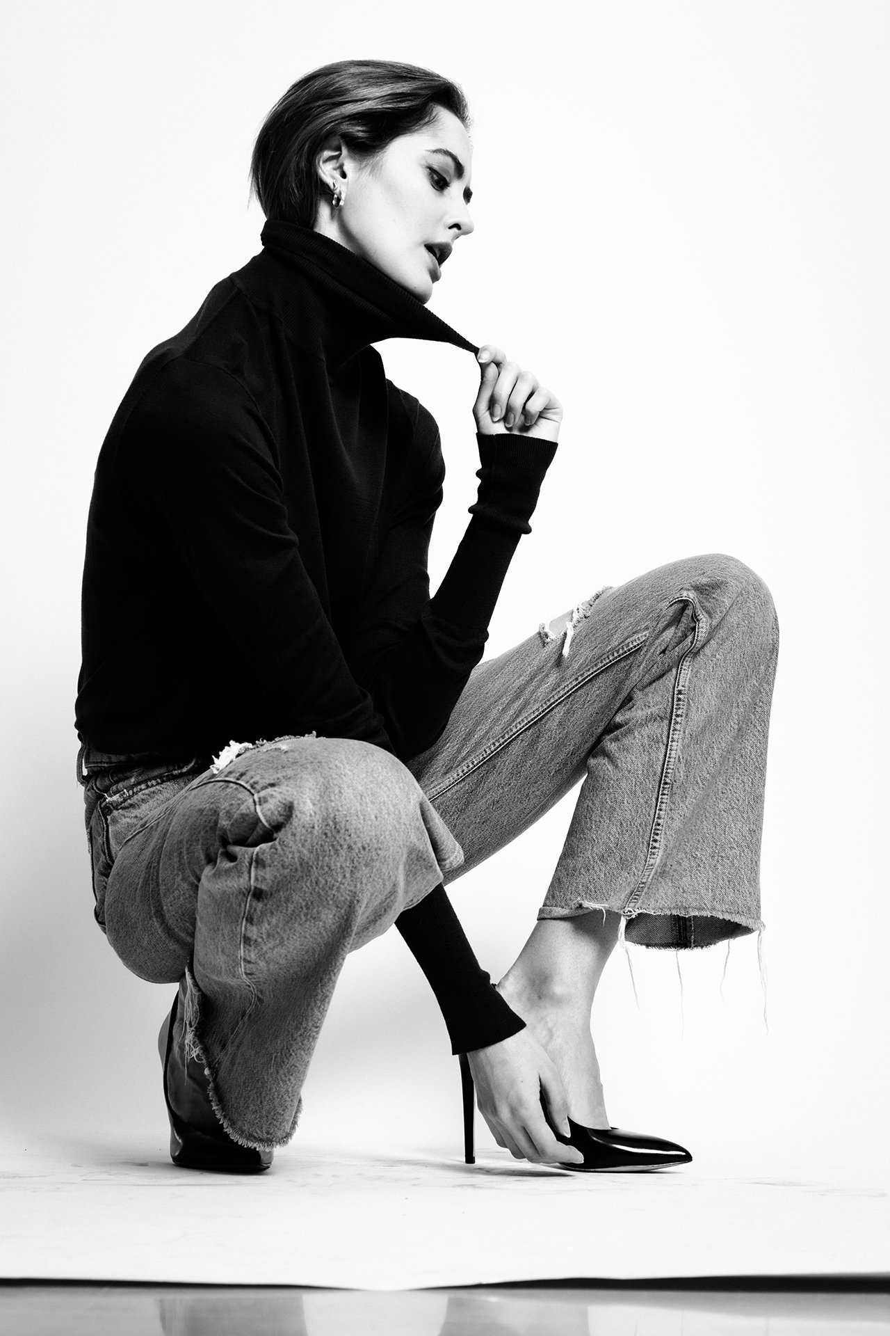 Black and white photo of a woman in a black turtleneck, ripped jeans, and high heels, sitting in a crouched position and looking down.