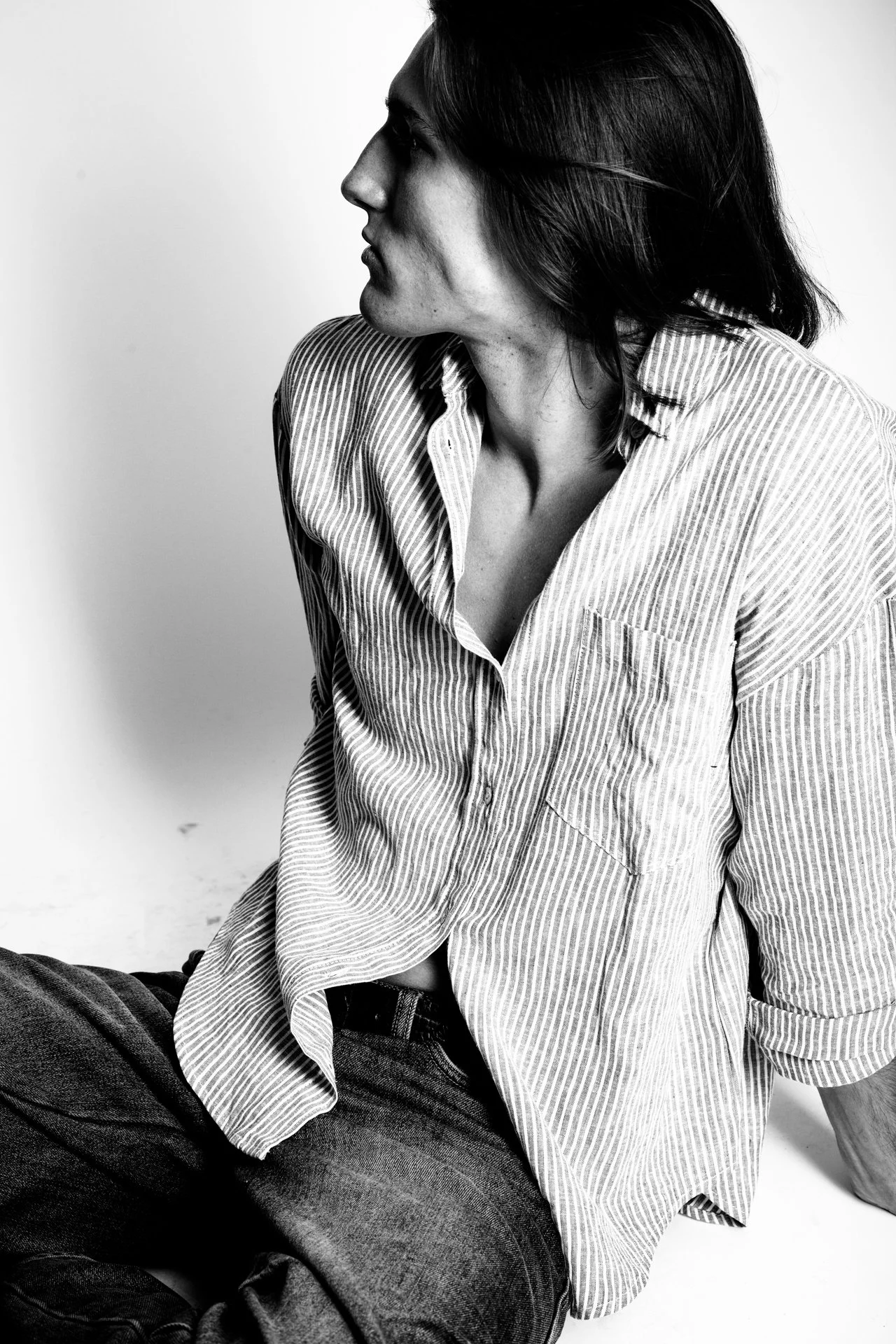 Black and white photo of a woman with medium-length dark hair, wearing a striped button-up shirt and jeans, sitting and looking to the left against a plain background.