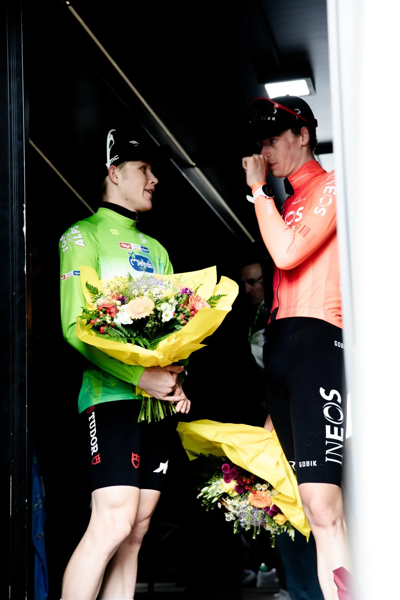 Two female cyclists in racing outfits having a conversation, one holding a bouquet of flowers, inside a dark room or trailer.