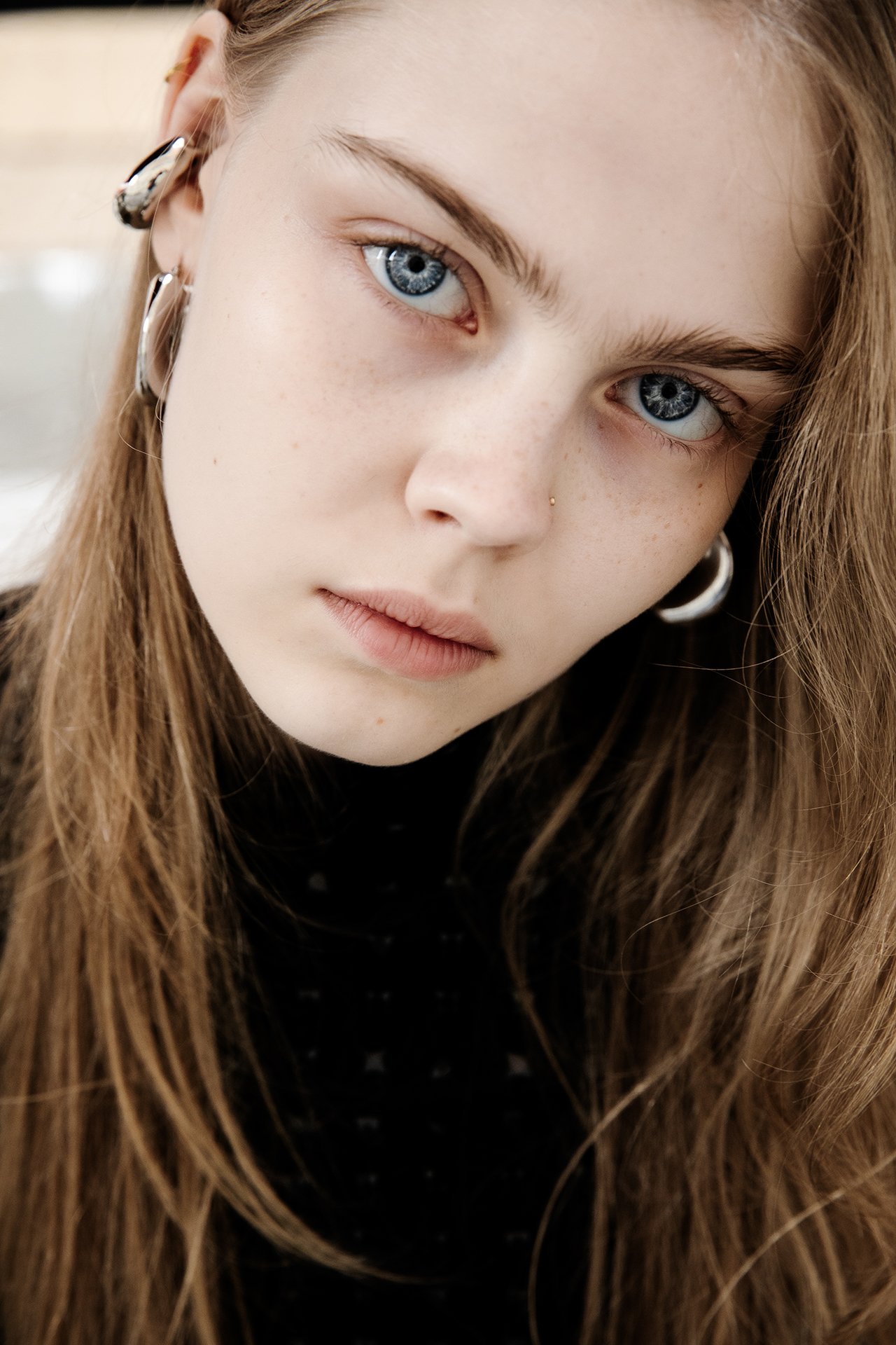 Close-up of a young woman with blue eyes, light skin, freckles, long brown hair, and silver hoop earrings, looking directly at the camera.
