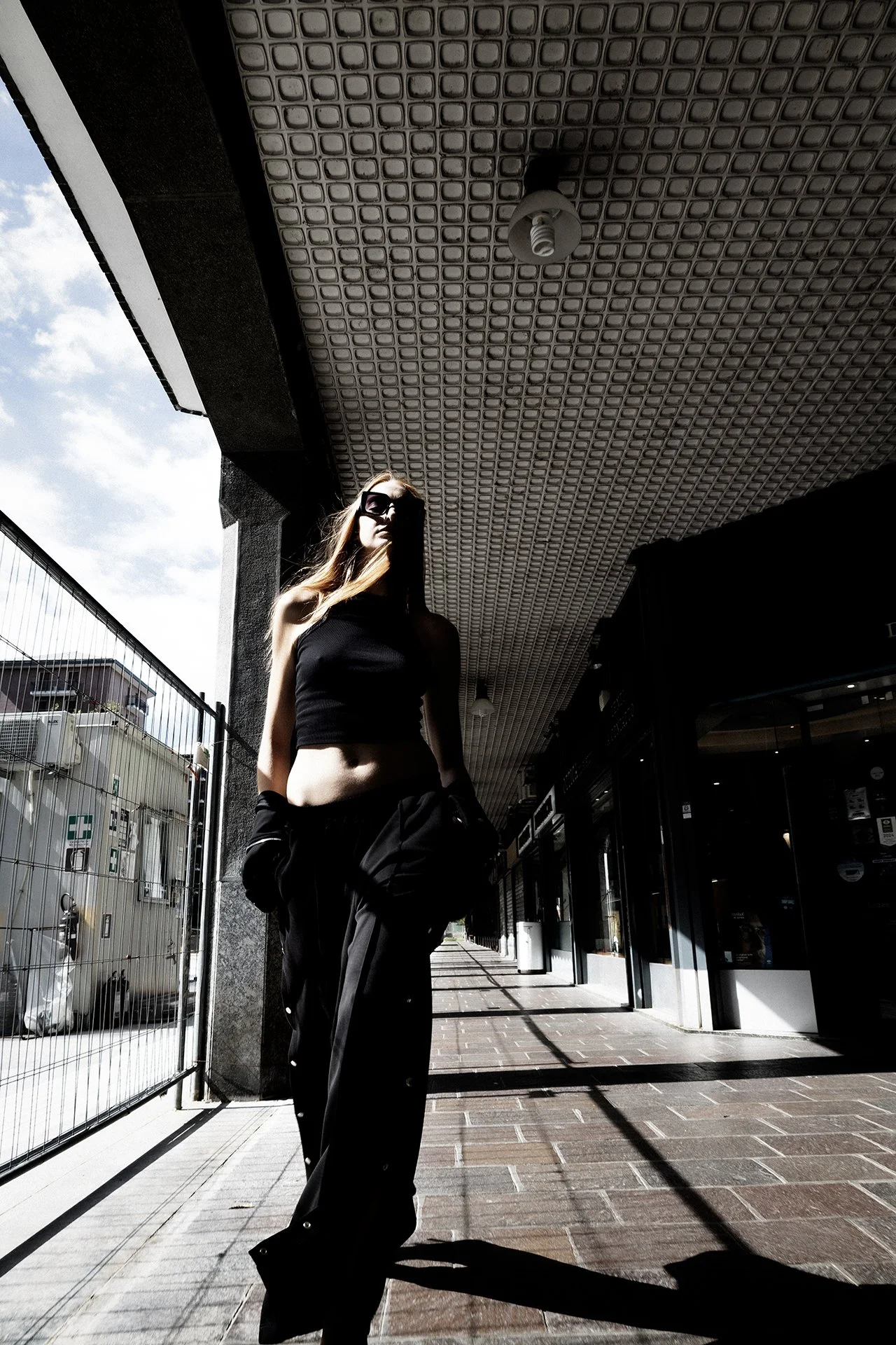 A woman wearing black clothing, including a crop top and pants, standing outside an urban shopping area with stores and a fence, under a patterned overhang, with the sun casting shadows.