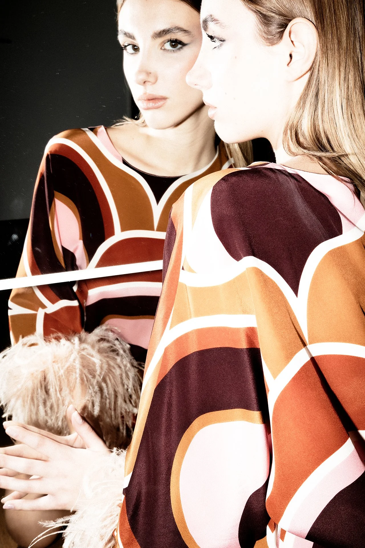 A woman looking into a mirror, wearing a colorful patterned dress with shades of brown, orange, pink, and black, with her reflection visible.