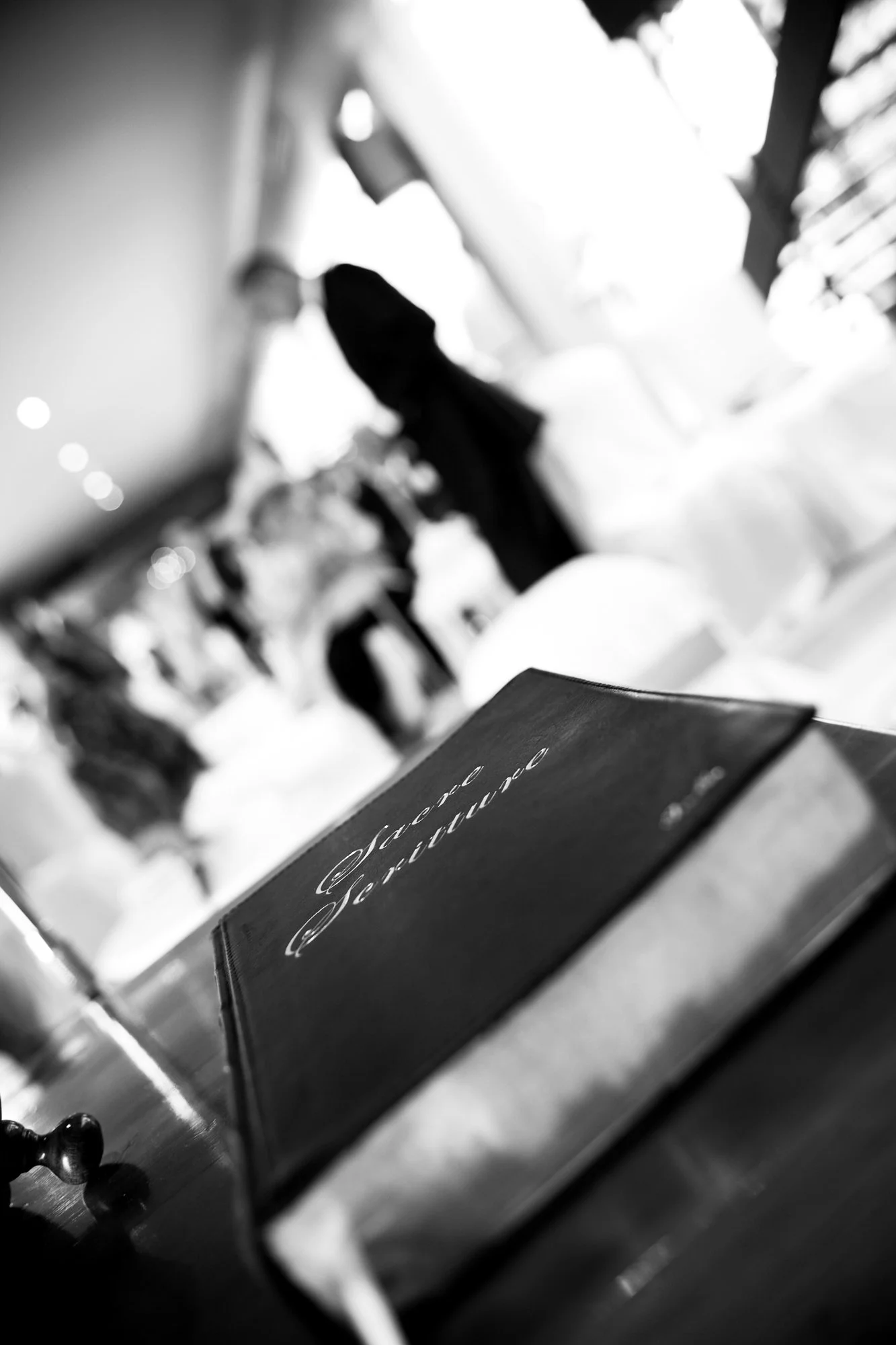 Black and white photo of a menu titled 'Menu de Servico' on a table at a restaurant or event, with blurred figures of people in the background.
