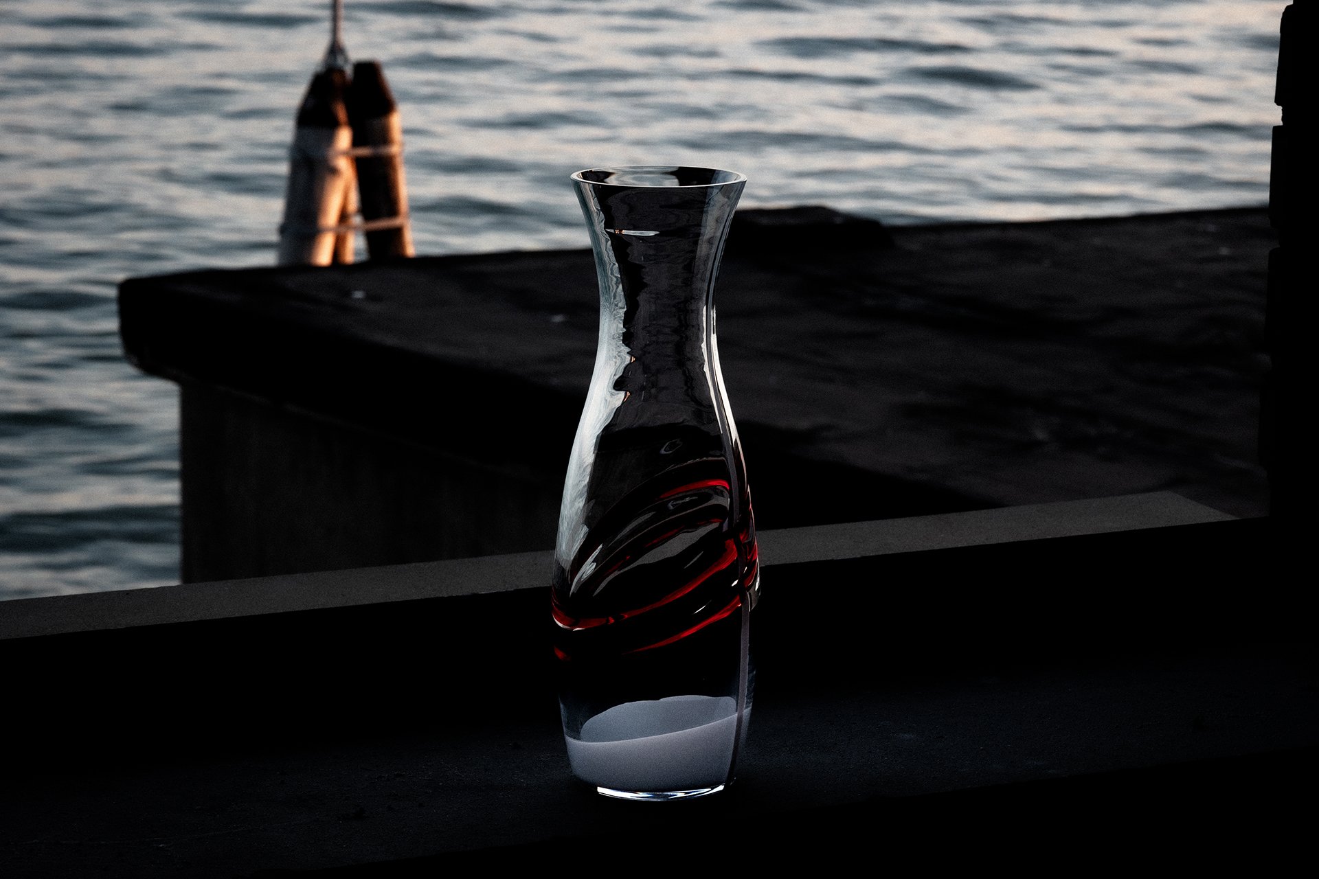 A glass vase with a decorative red swirl, partly filled with a white liquid, sits on a dark surface near a body of water at sunset.