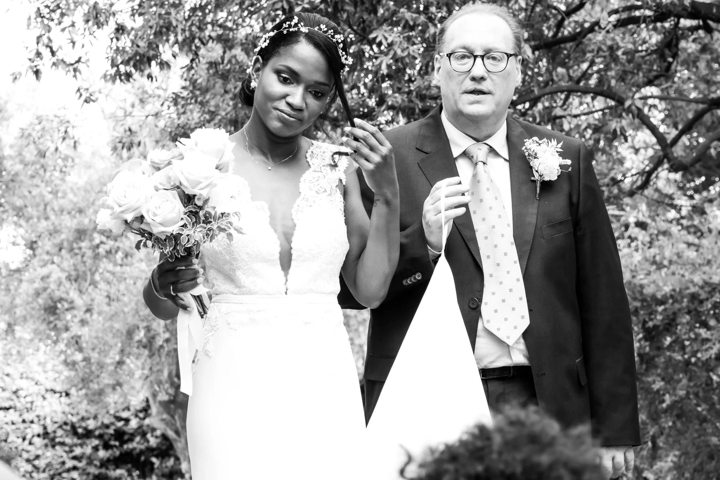A black-and-white photo of a bride and an older man, possibly her father, at a wedding ceremony outdoors. The bride holds a bouquet of roses and has a crown of flowers in her hair. The older man wears glasses, a suit, and a tie, with a boutonniere on