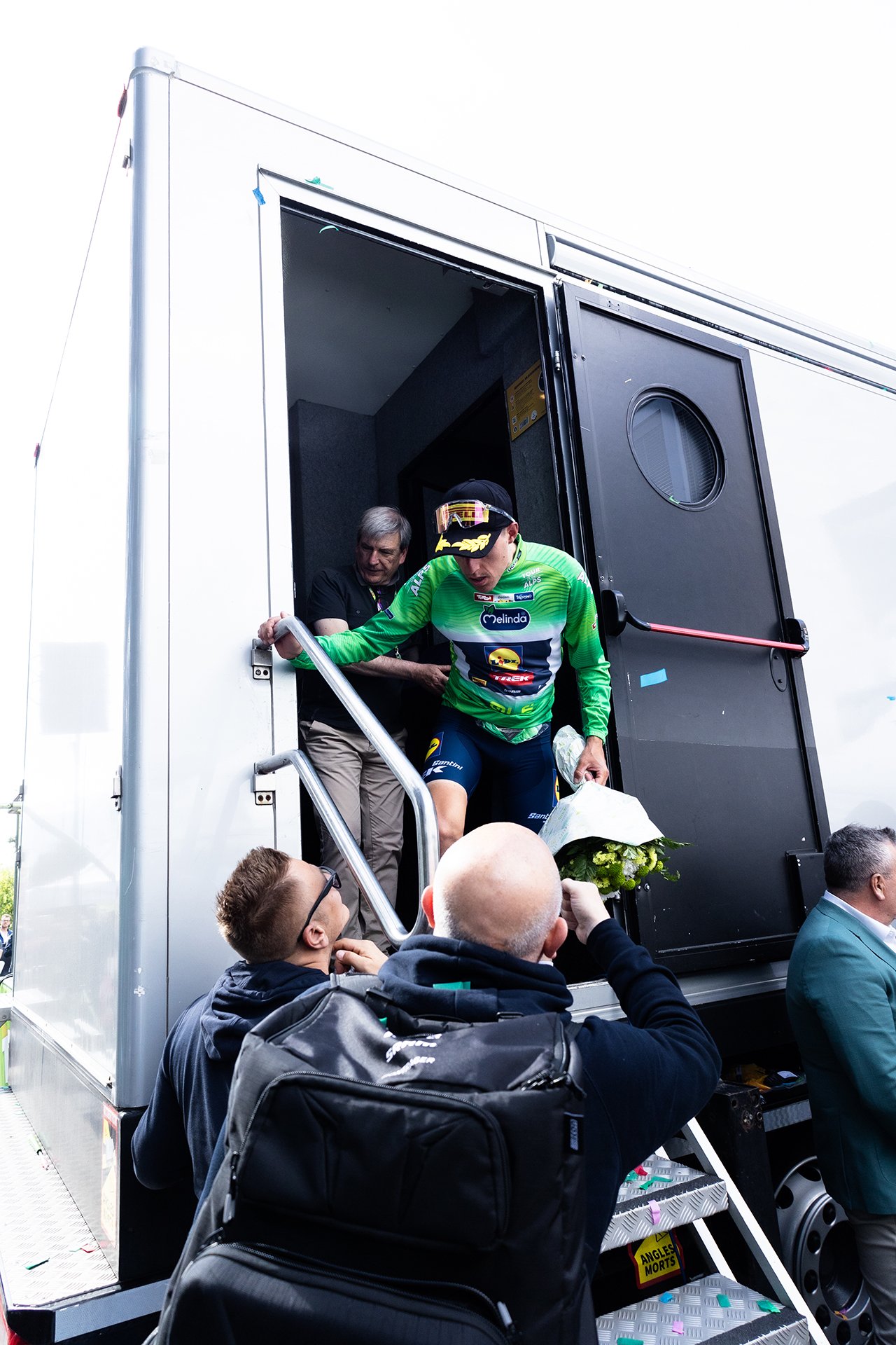 Cyclist exiting a mobile podium, holding a bouquet of flowers, shakes hands with a man at the top of the stairs, while several people watch.