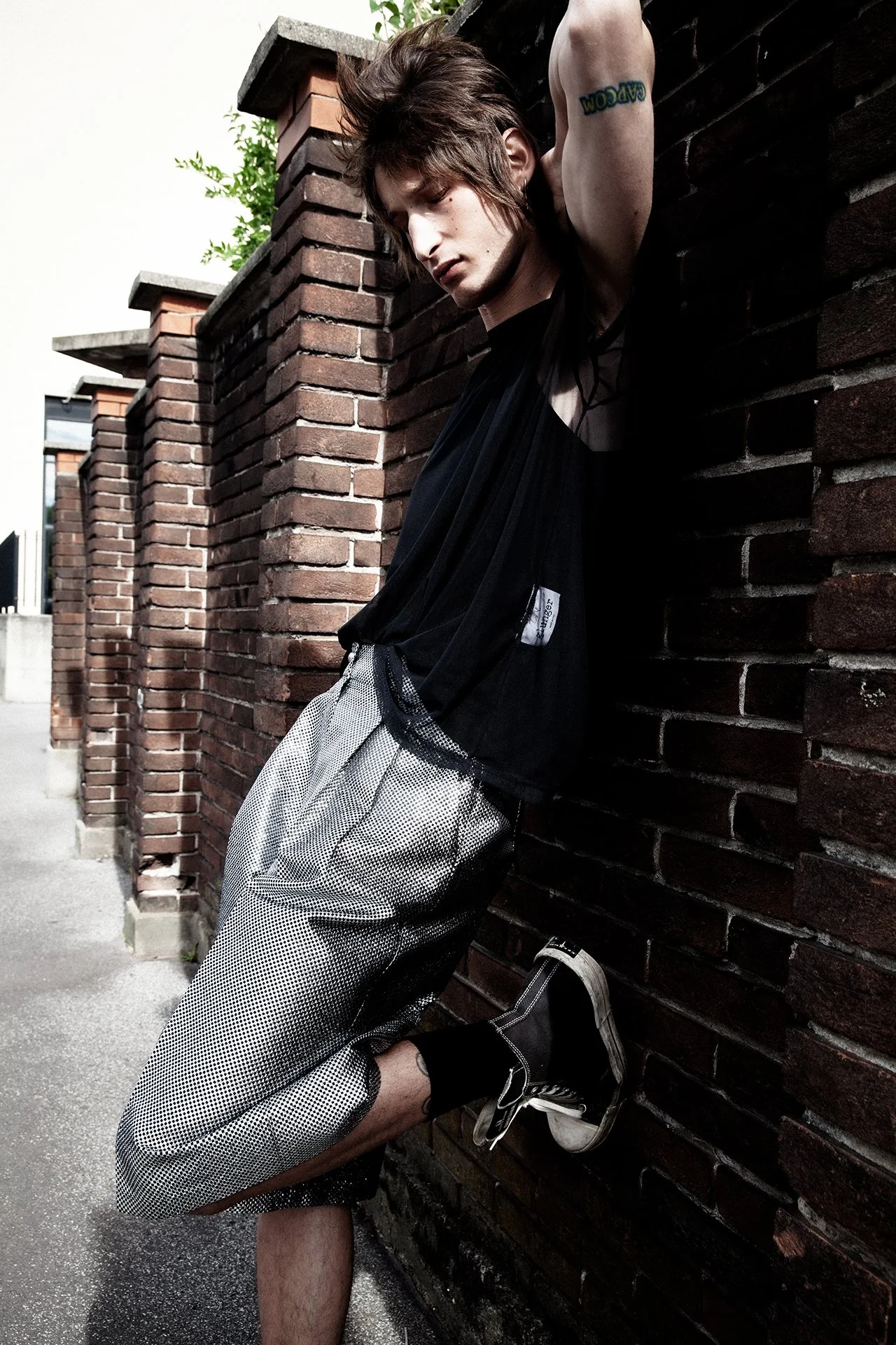 A young man with tousled brown hair, leaning against a brick wall with one arm raised above his head. He is wearing a black sleeveless mesh shirt, patterned shorts, black socks, and black sneakers. The setting appears to be outdoors, with a concrete 