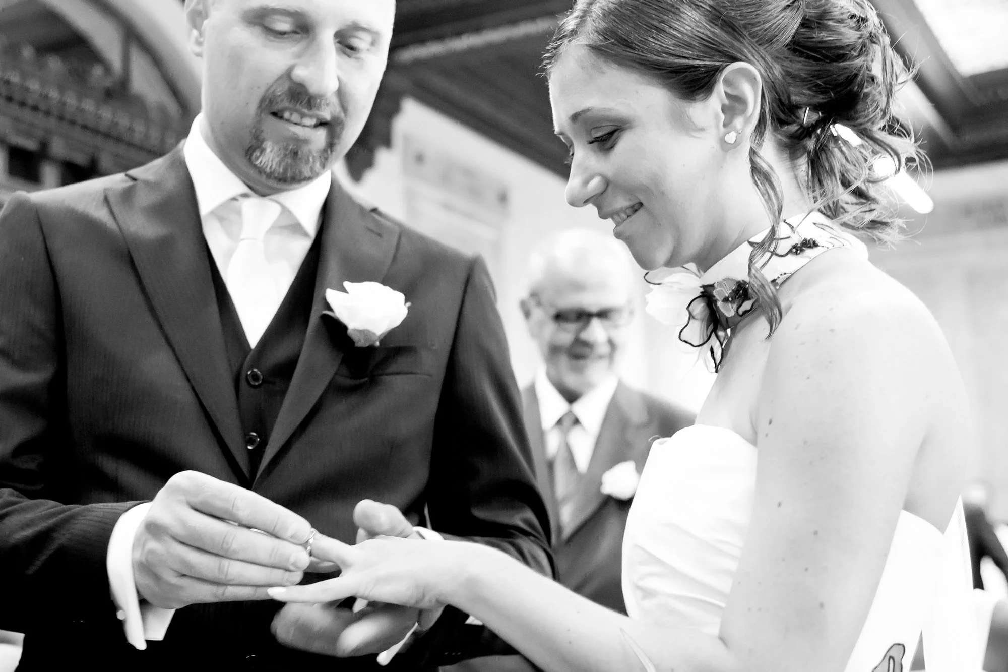 A black and white photo of a man and woman during a wedding ceremony. The man is placing a ring on the woman's finger. The man is wearing a suit with a boutonnière, and the woman is dressed in a wedding gown with a floral necklace. They are smiling a