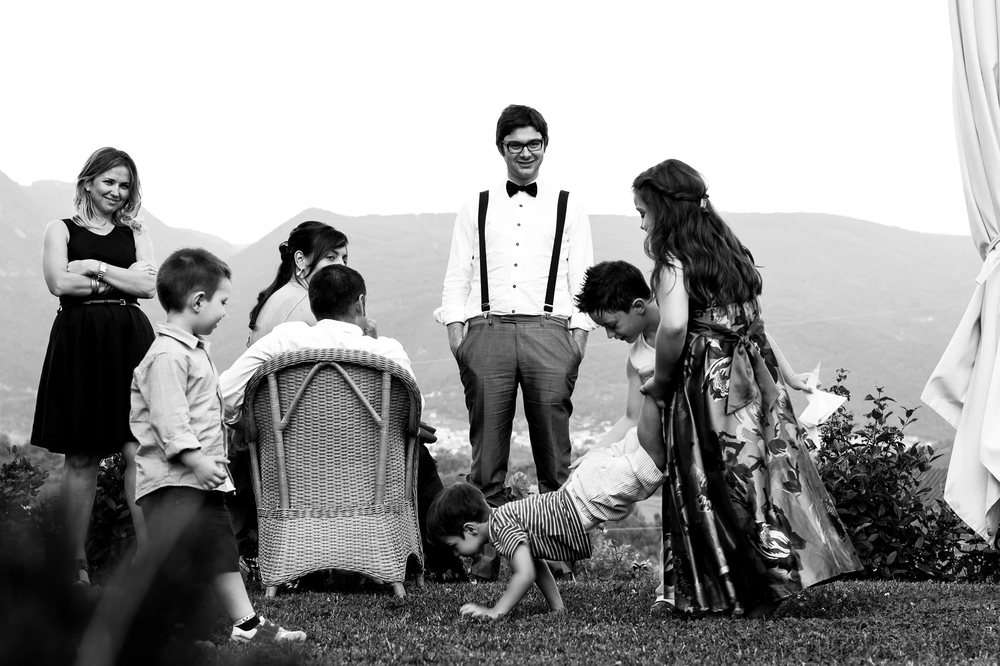 Group of children and an adult man outdoors in a scenic mountainous area, with some children playing and others watching, in black and white.