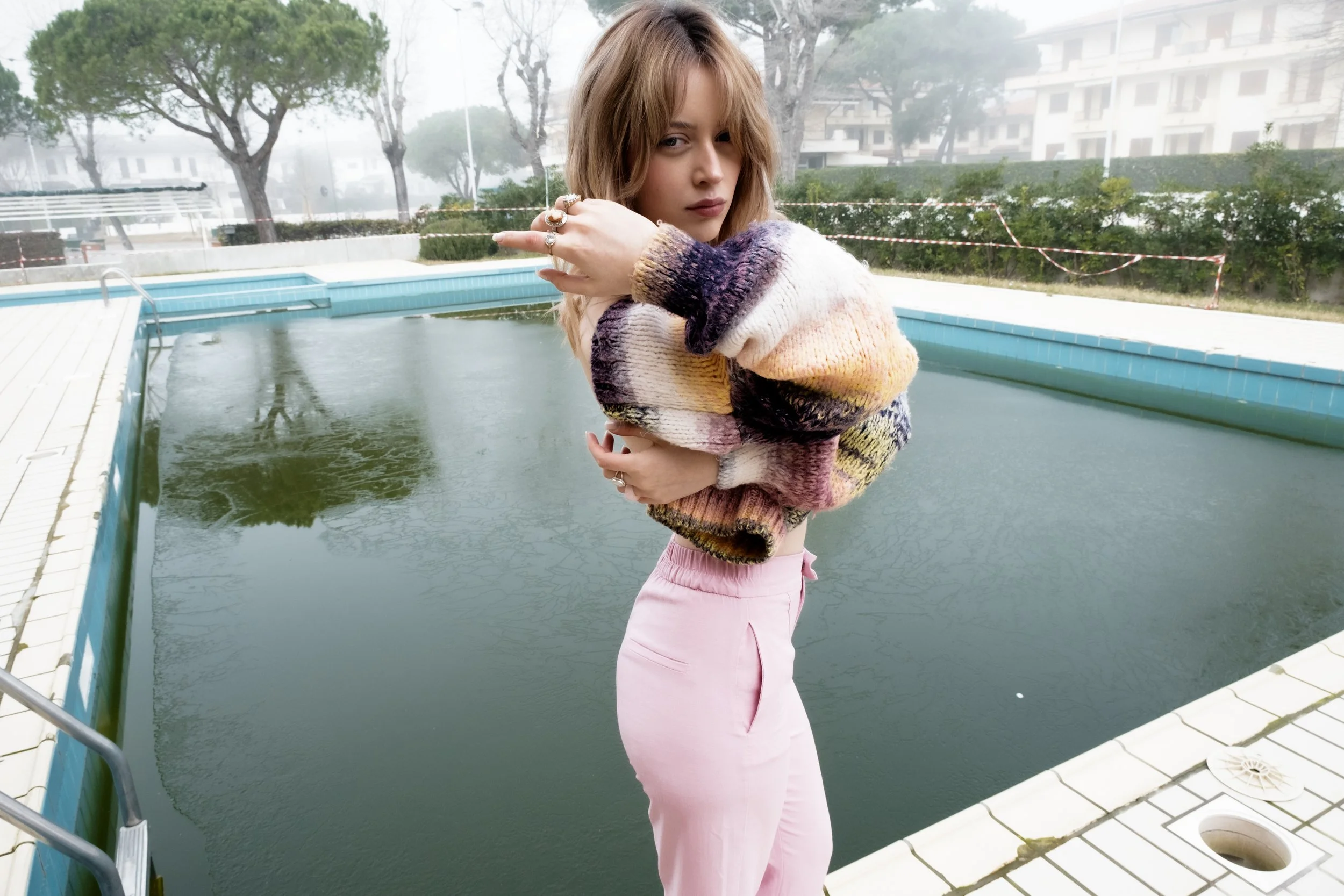 A young woman in pastel pink pants and a colorful knitted sweater stands next to an empty outdoor pool with foggy trees and buildings in the background.