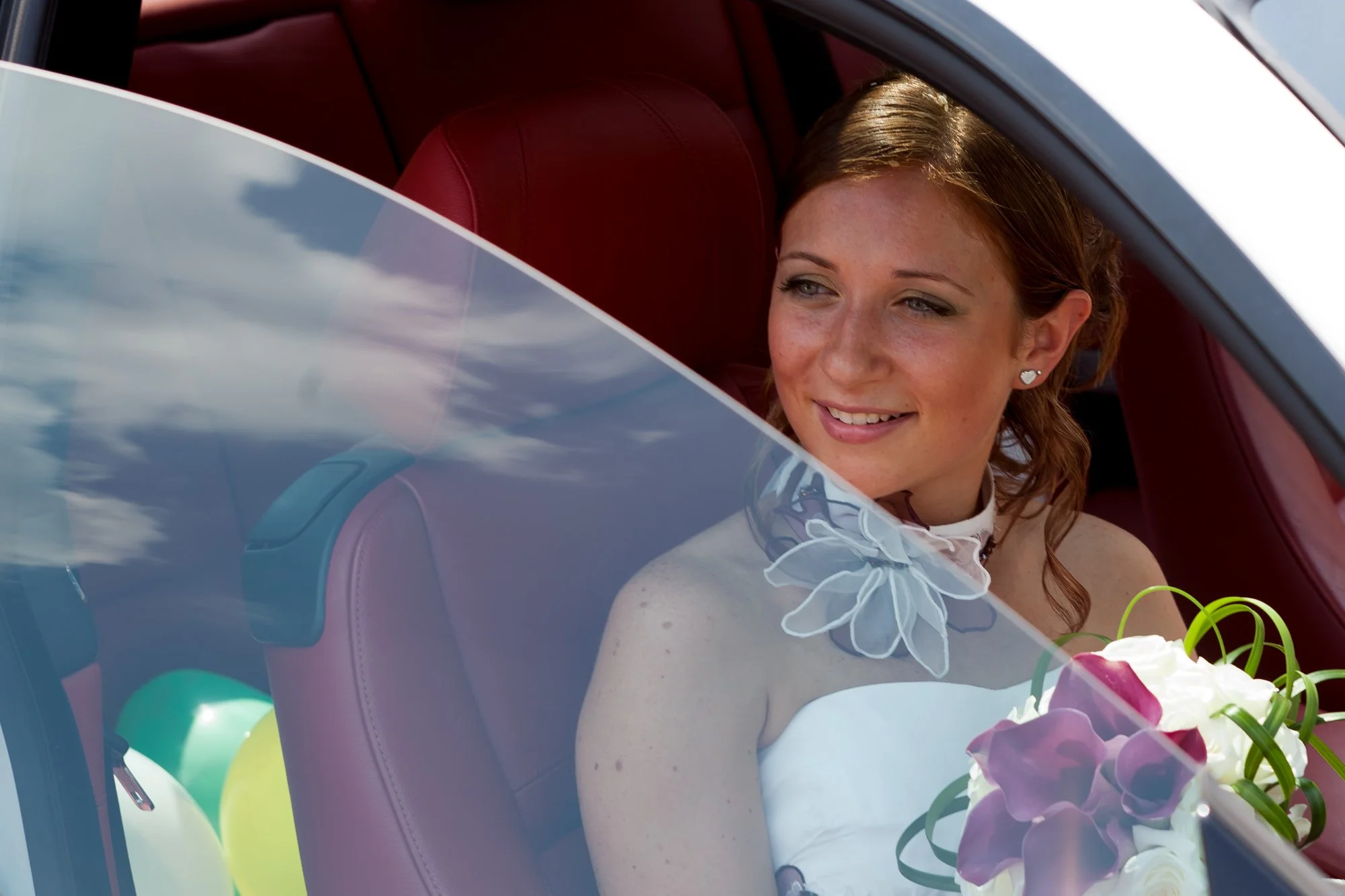 A woman with auburn hair and heart-shaped earrings sitting inside a car, smiling and holding a bouquet of flowers.