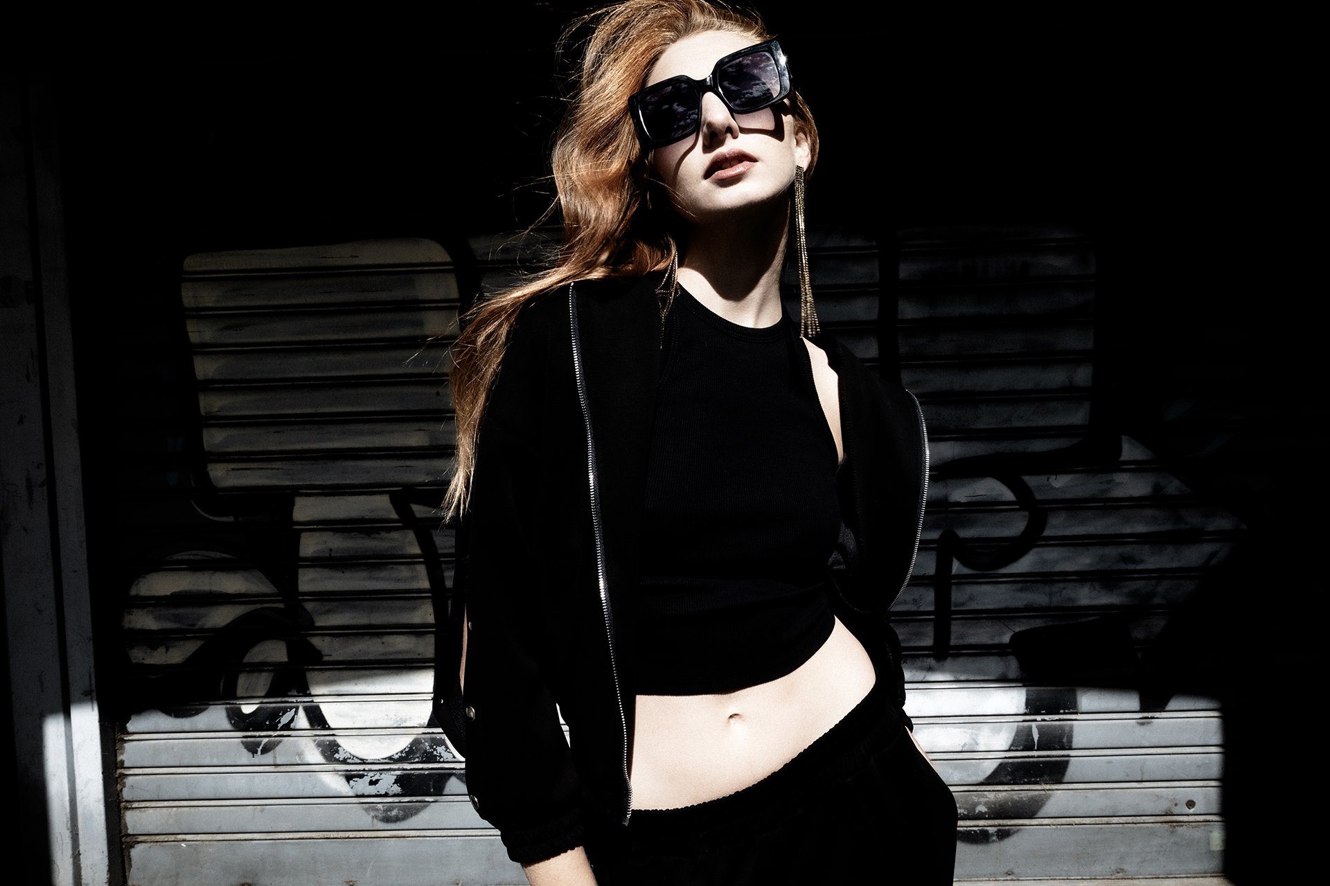 A woman with long red hair wearing large black sunglasses, a black crop top, black pants, and a black jacket, standing in front of a graffiti-covered metal shutter in bright sunlight.