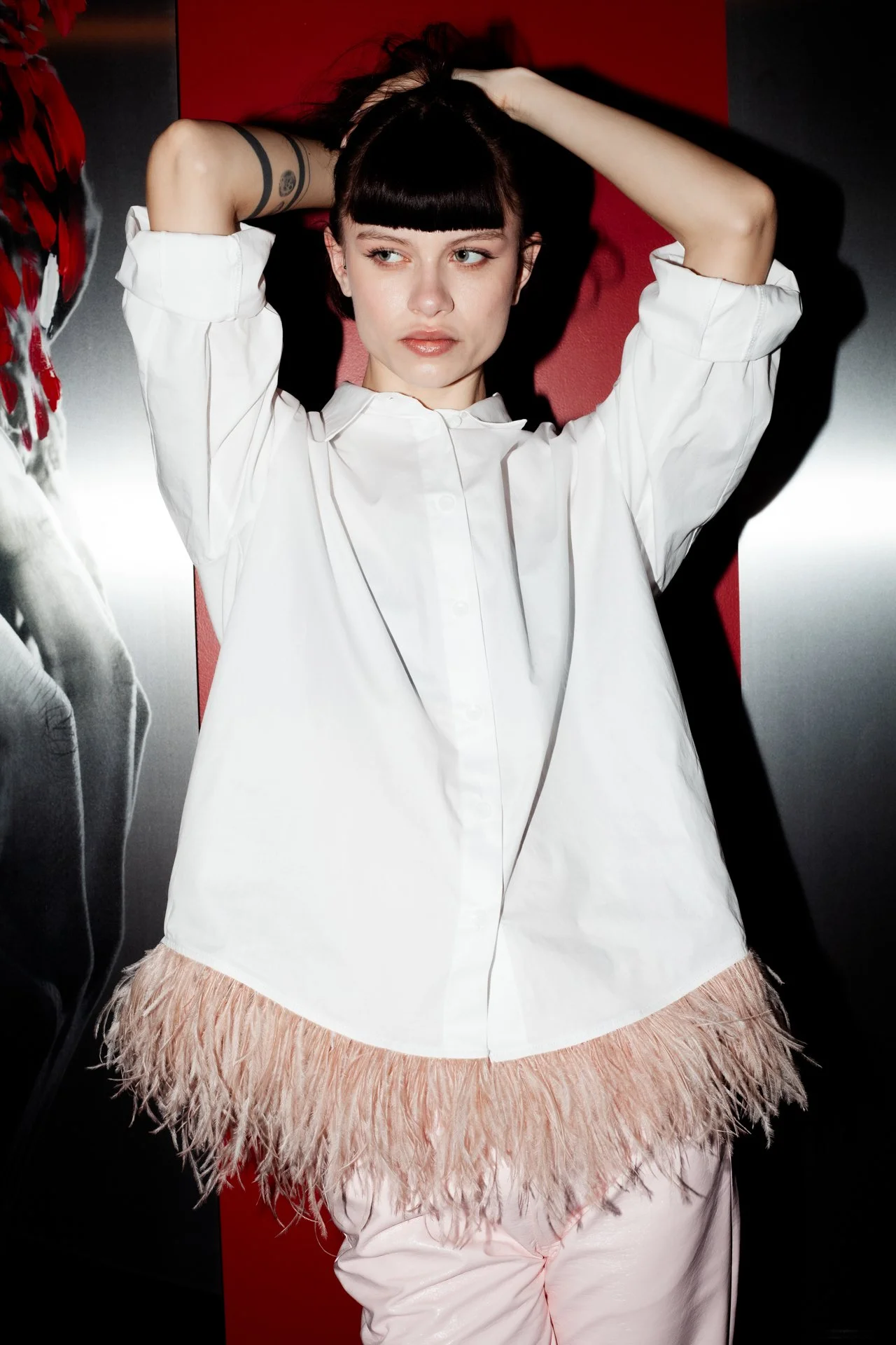 A young woman with dark hair and blunt bangs, wearing a white button-up shirt with feathered trim at the bottom, posing with her hands behind her head against a red and gray background.