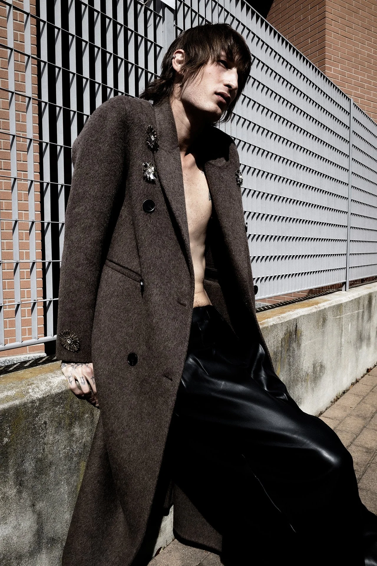 A young man with brown hair, wearing an open brown overcoat with decorative embellishments, black leather pants, and no shirt, leaning against a concrete ledge outdoors.