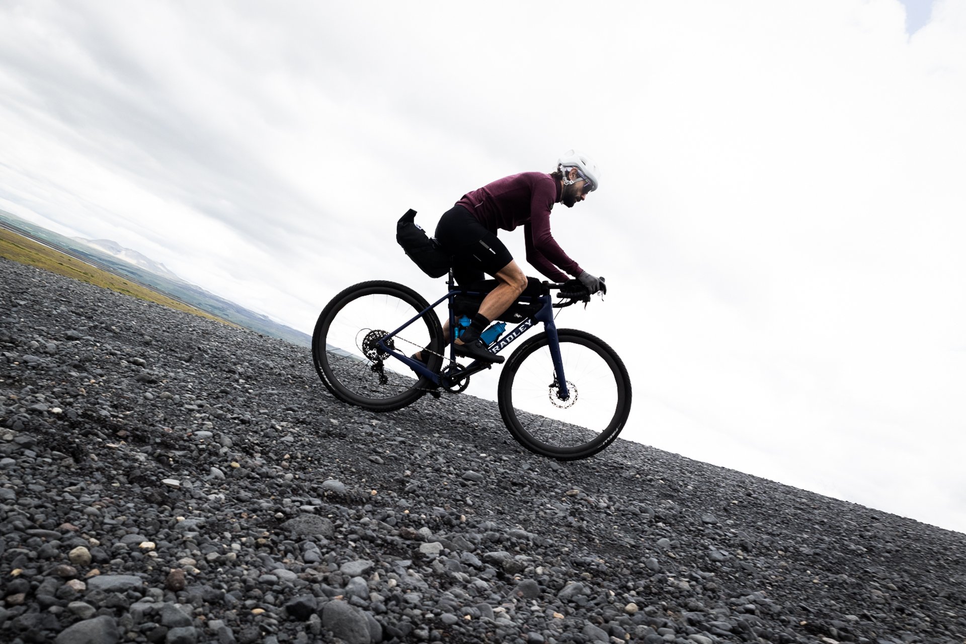 Cyclist wearing a helmet and maroon sweatshirt riding a mountain bike on a rocky terrain under a cloudy sky.