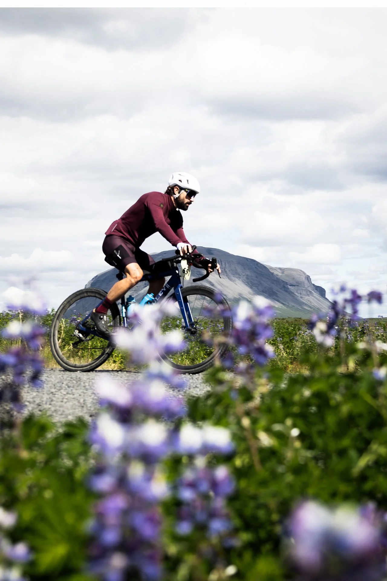 A man riding a mountain bike on a trail with mountains and a partly cloudy sky in the background, and purple flowers in the foreground.