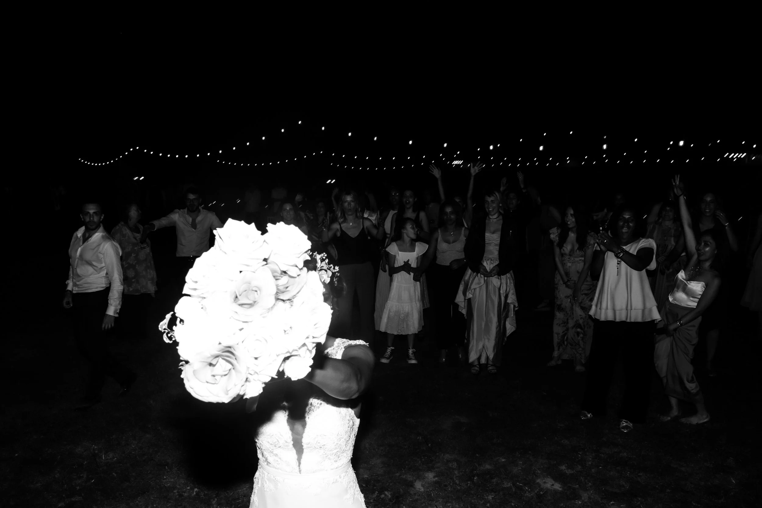A bride holding a bouquet of flowers in front of a large group of people during a nighttime celebration with string lights overhead.