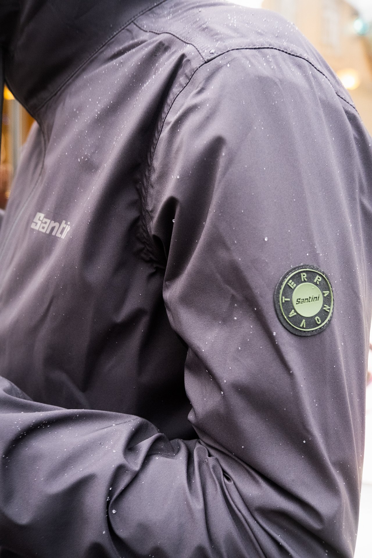 Close-up of a dark gray Santini rain jacket with a circular Santini badge on the sleeve, with small water droplets on the fabric.