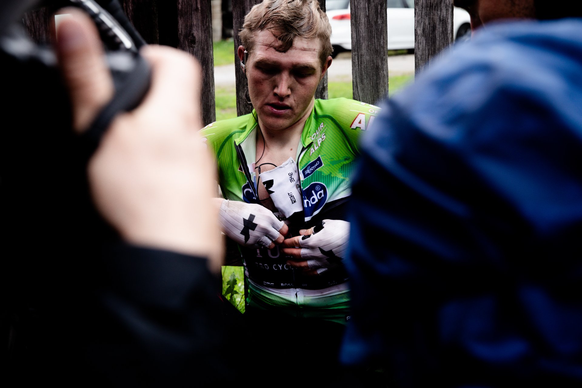 A male cyclist in green and black gear getting assistance from team members after a race, with a fence and parked cars in the background.