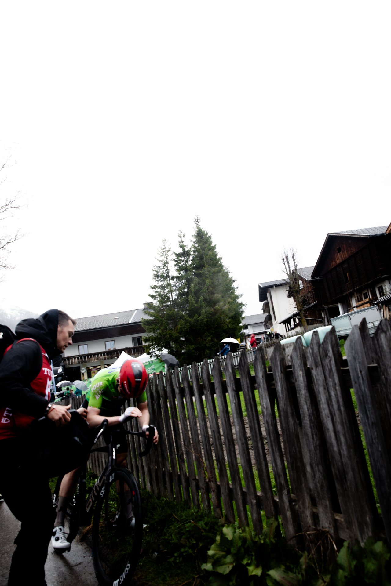 A cyclist in a green jersey and red helmet leaning over his bike, being assisted by a man in a black jacket, near a wooden fence in a residential area on a rainy day, with spectators holding umbrellas in the background.
