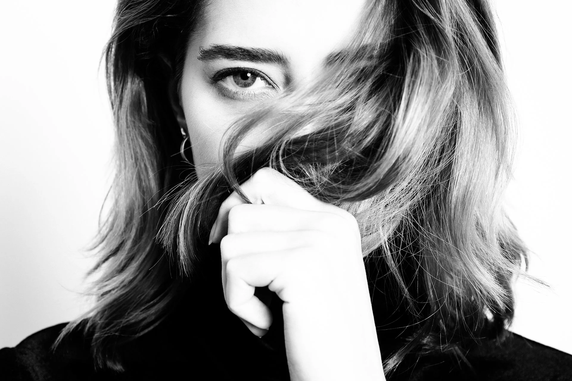 Black and white portrait of a woman with wavy hair covering part of her face, one eye visible and looking at the camera, with her hand near her chin.