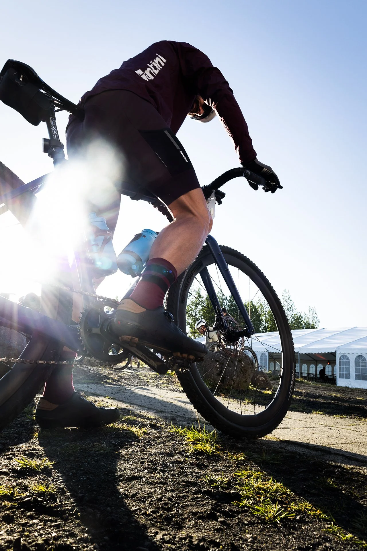 A person riding a mountain bike outdoors with sunlight shining behind, wearing cycling gear and helmet, on a dirt path near a white event tent.