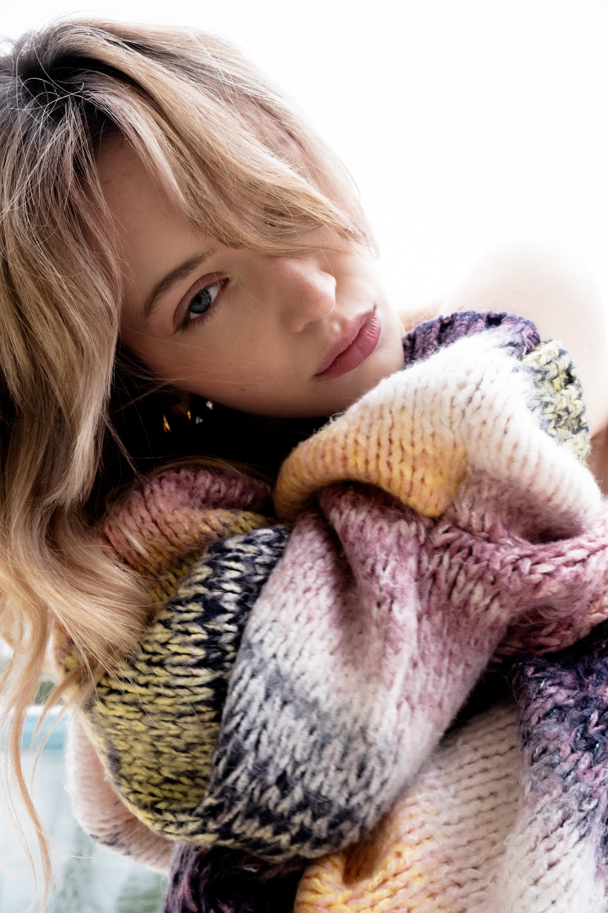 Close-up of a woman with blonde hair wearing a multicolored knitted sweater, with a neutral expression, indoors with a white background.