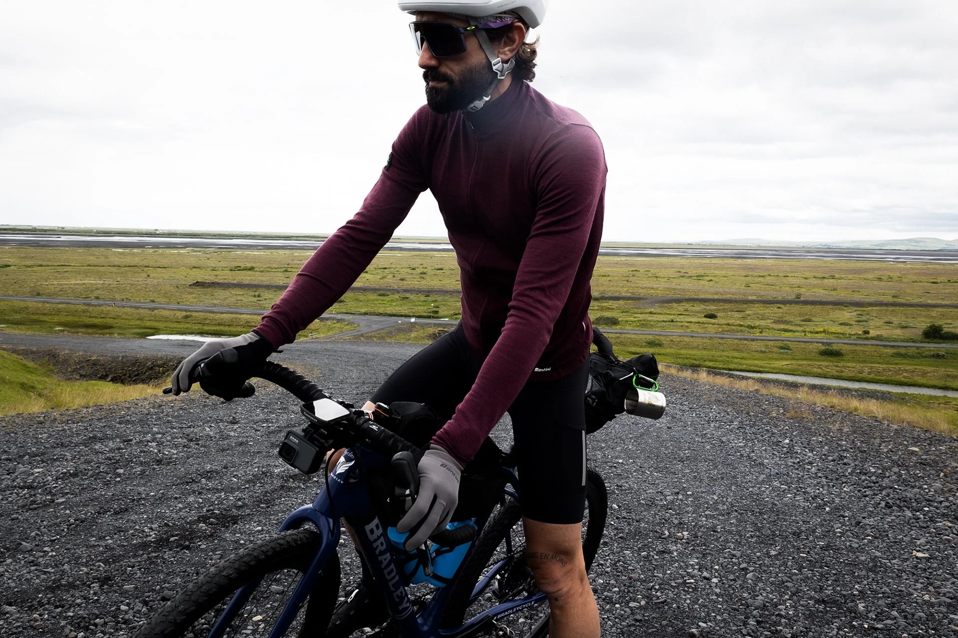 A man wearing a maroon long-sleeve shirt, black shorts, sunglasses, helmet, and gloves riding a blue mountain bike on a gravel path in an open grassy landscape with cloudy skies.