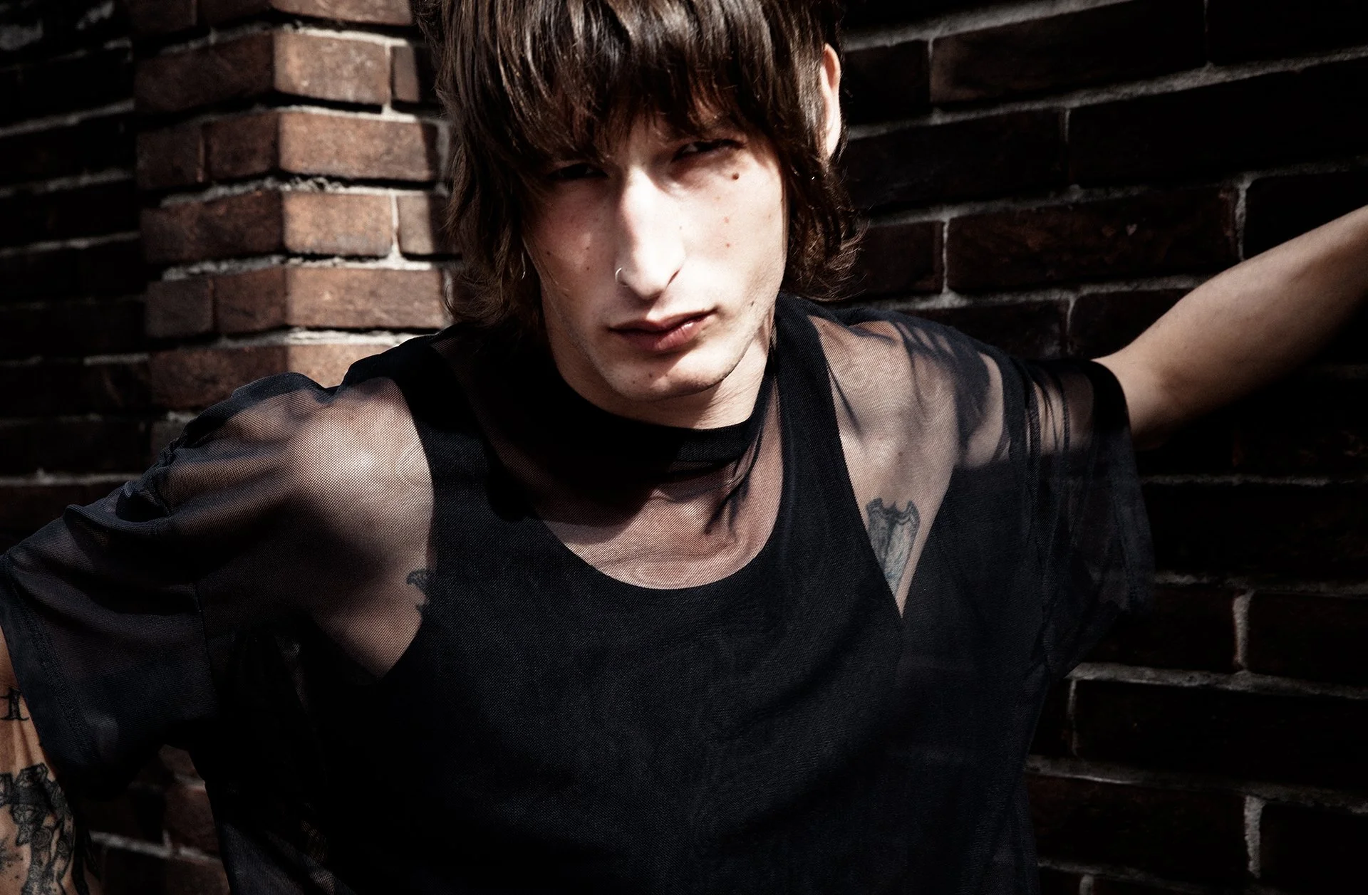 Young person with dark hair, wearing a black sheer shirt, standing against a brick wall with tattoos visible on their arm and chest.