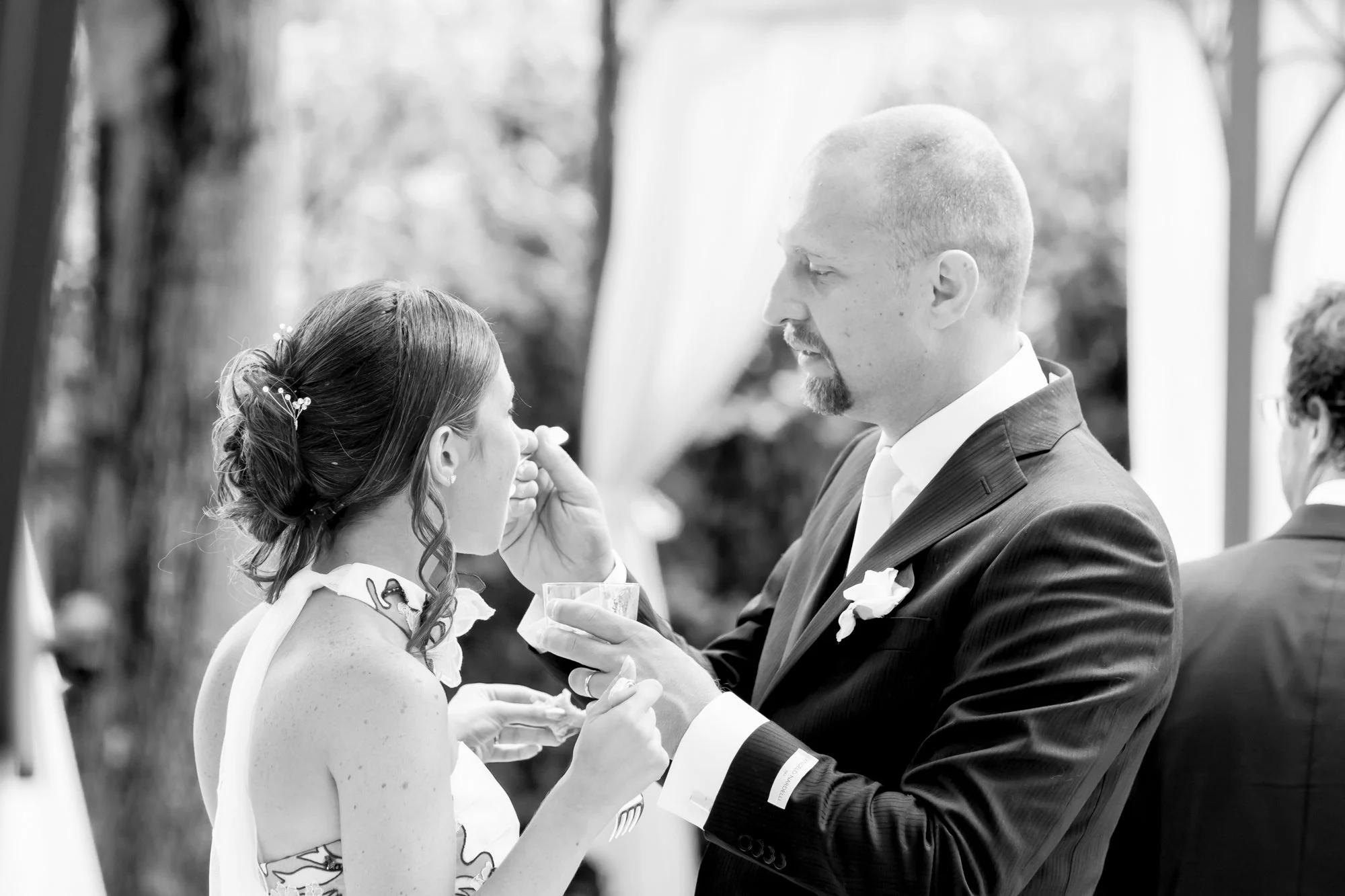 A groom with a beard and suit touches the bride's face during a wedding ceremony outdoors, with another man in the background.
