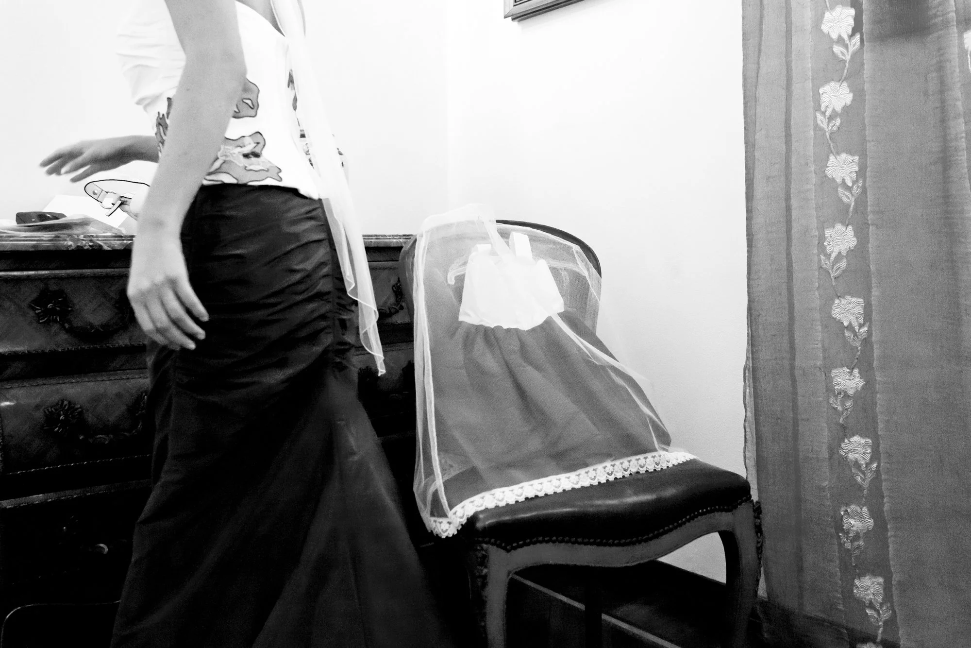 Part of a person in a dark skirt standing next to a wooden dresser and a chair with a lace veil on it, in a room with a curtain.
