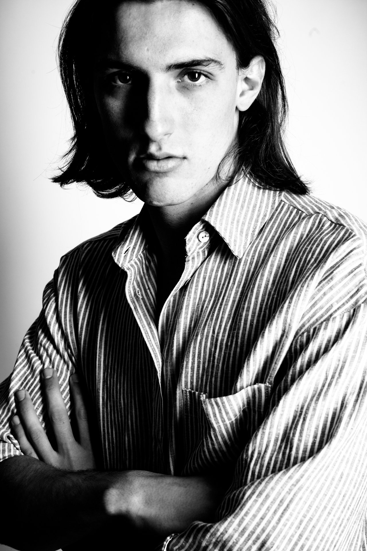 Black and white portrait of a young man with long dark hair wearing a striped button-up shirt with arms crossed.