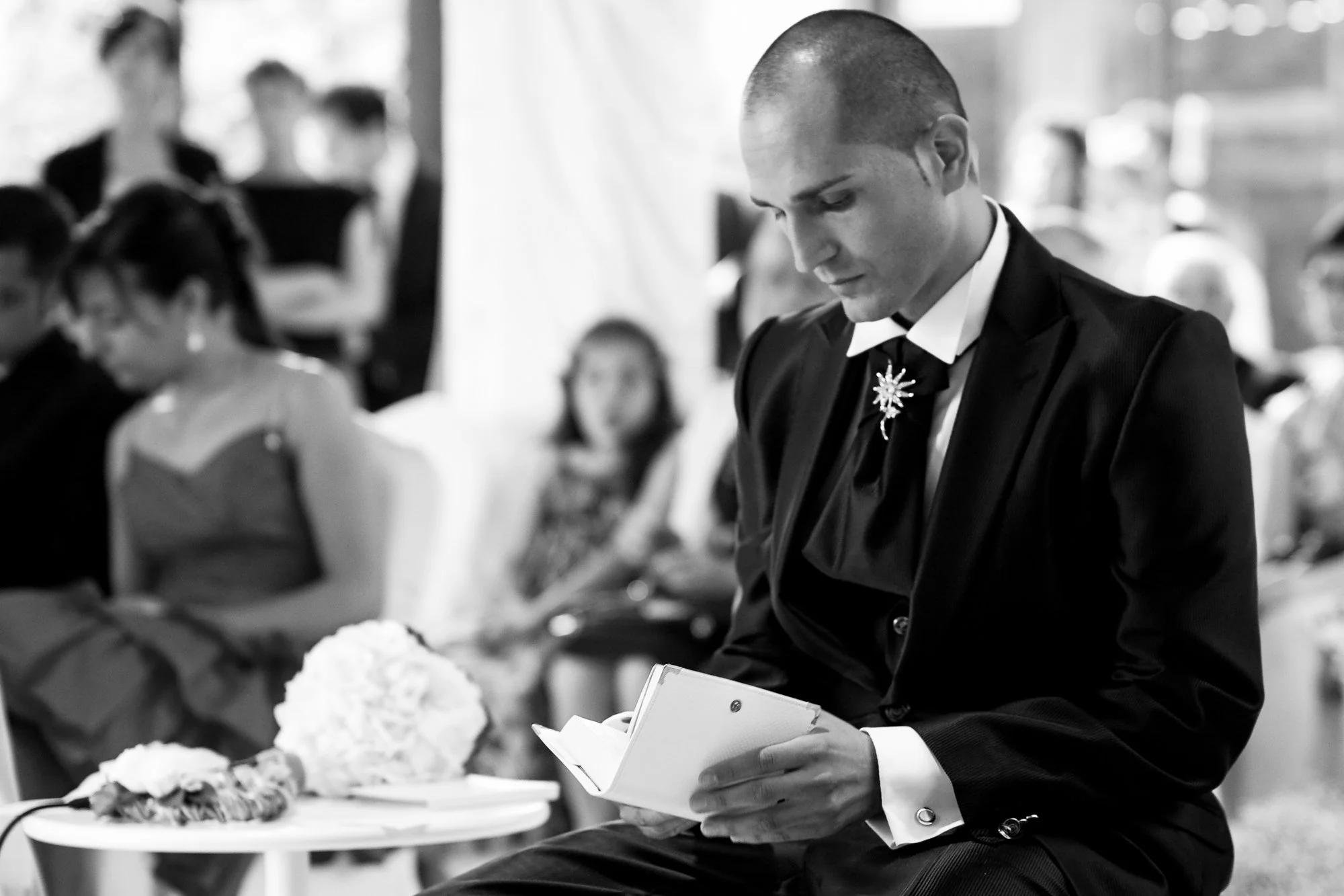 A man in a tuxedo with a flower lapel pin is reading from a notebook during a formal event, with seated guests in the background.