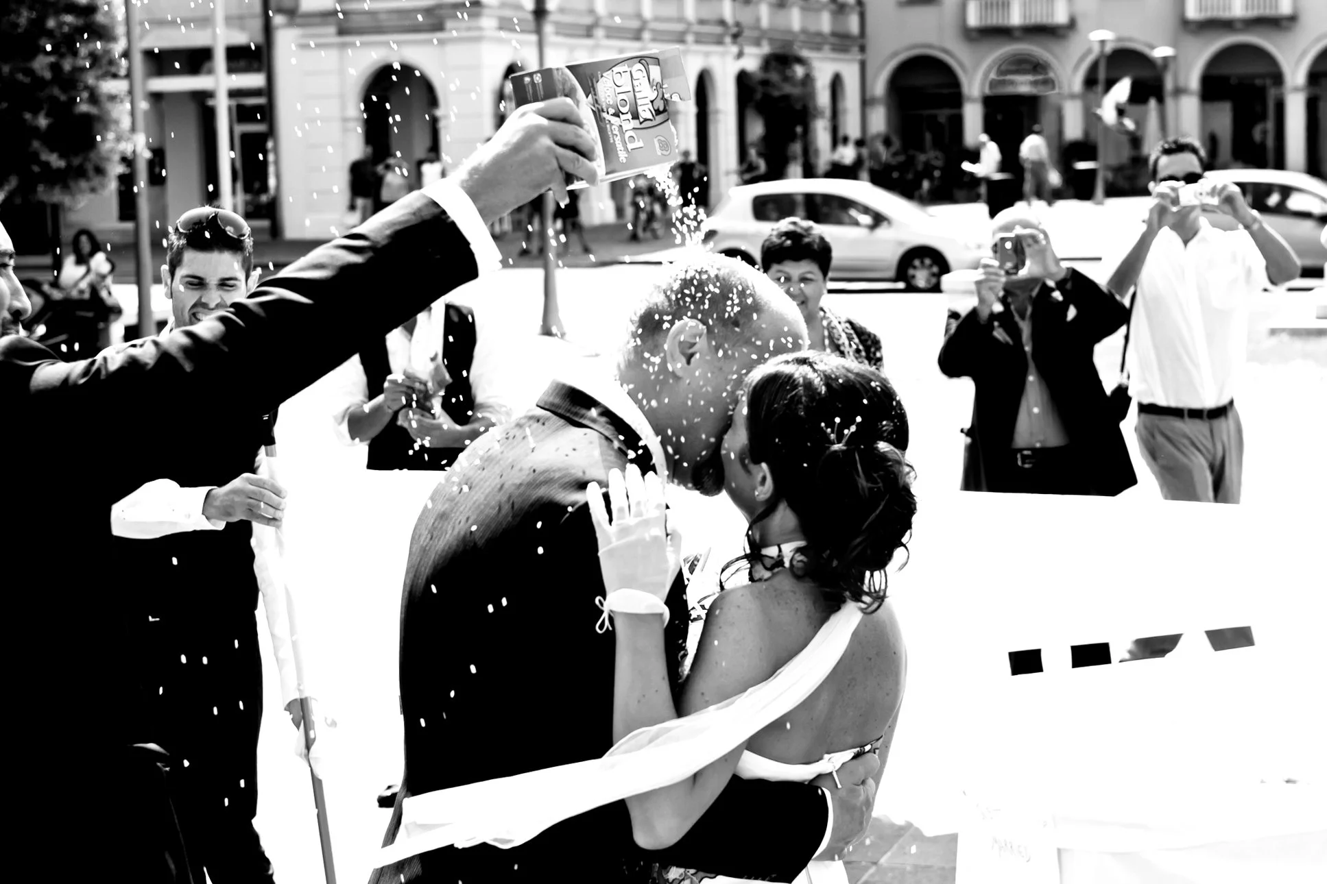 Black and white photo of a wedding couple kissing, surrounded by friends and family taking photos, with confetti falling around them.