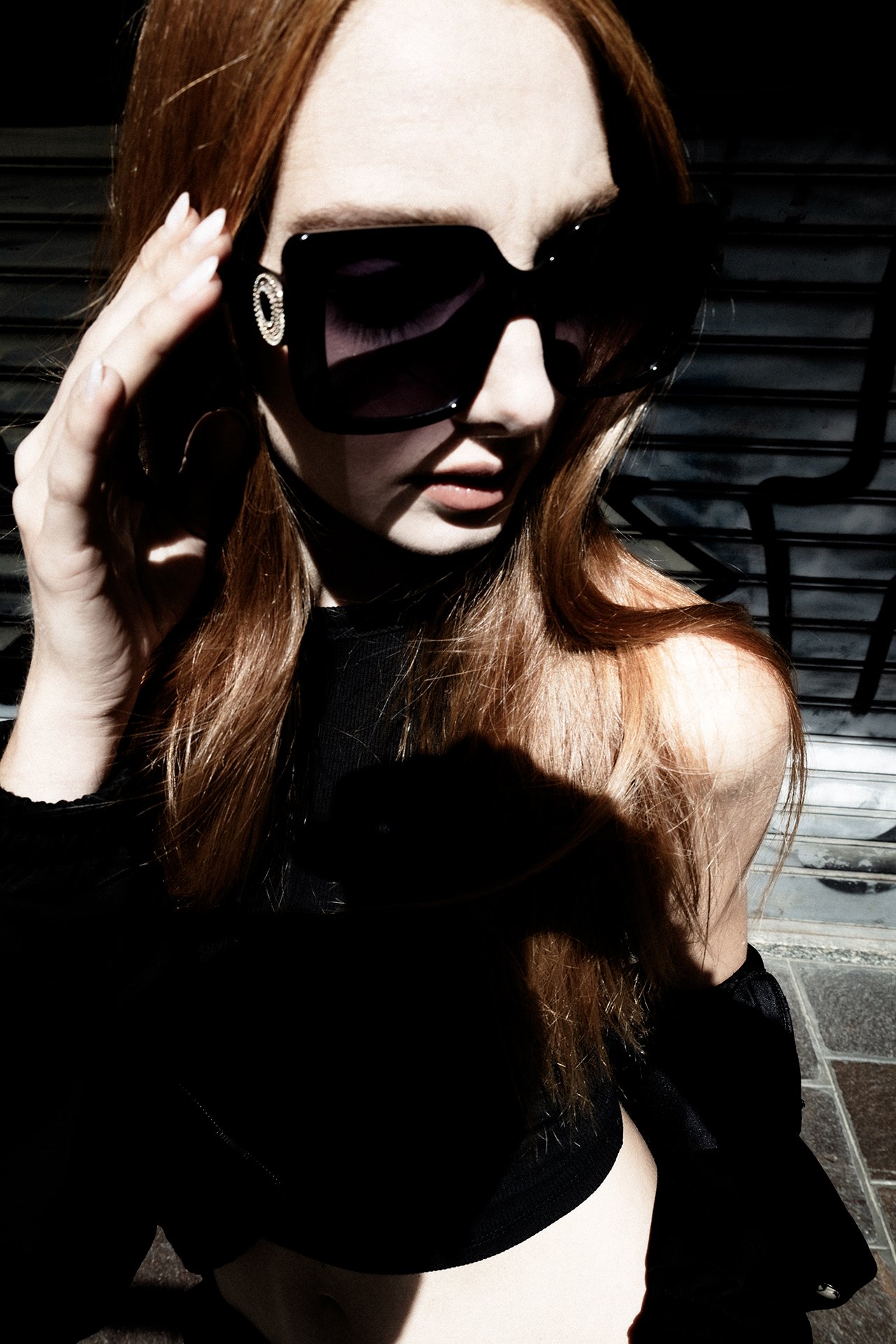 A woman with red hair wearing large black sunglasses, earrings, and a black outfit, standing outside with sunlight and shadows creating dramatic effects.