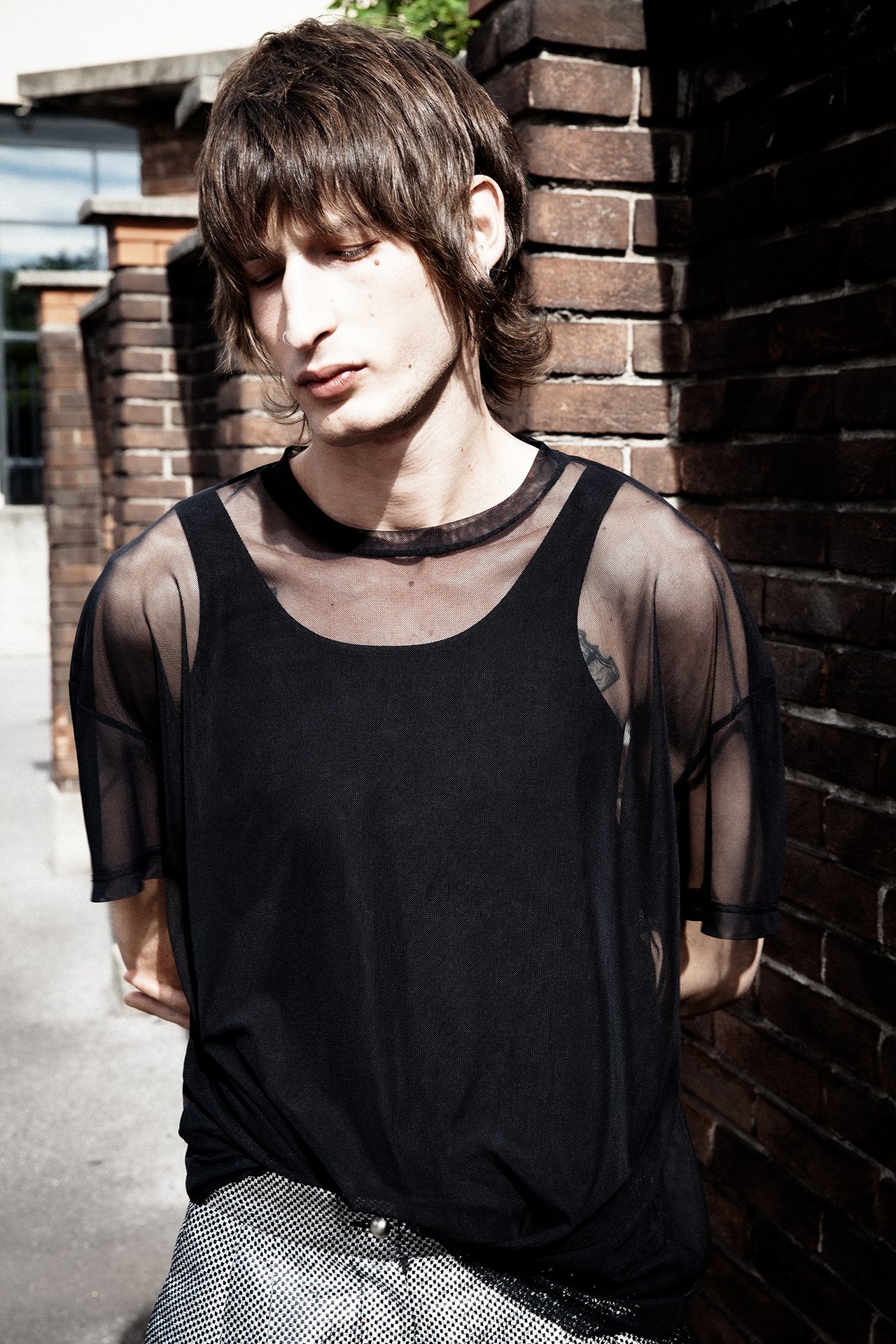 A young man with brown, shaggy hair standing against a brick wall outdoors, wearing a black sheer top with a black tank top underneath.