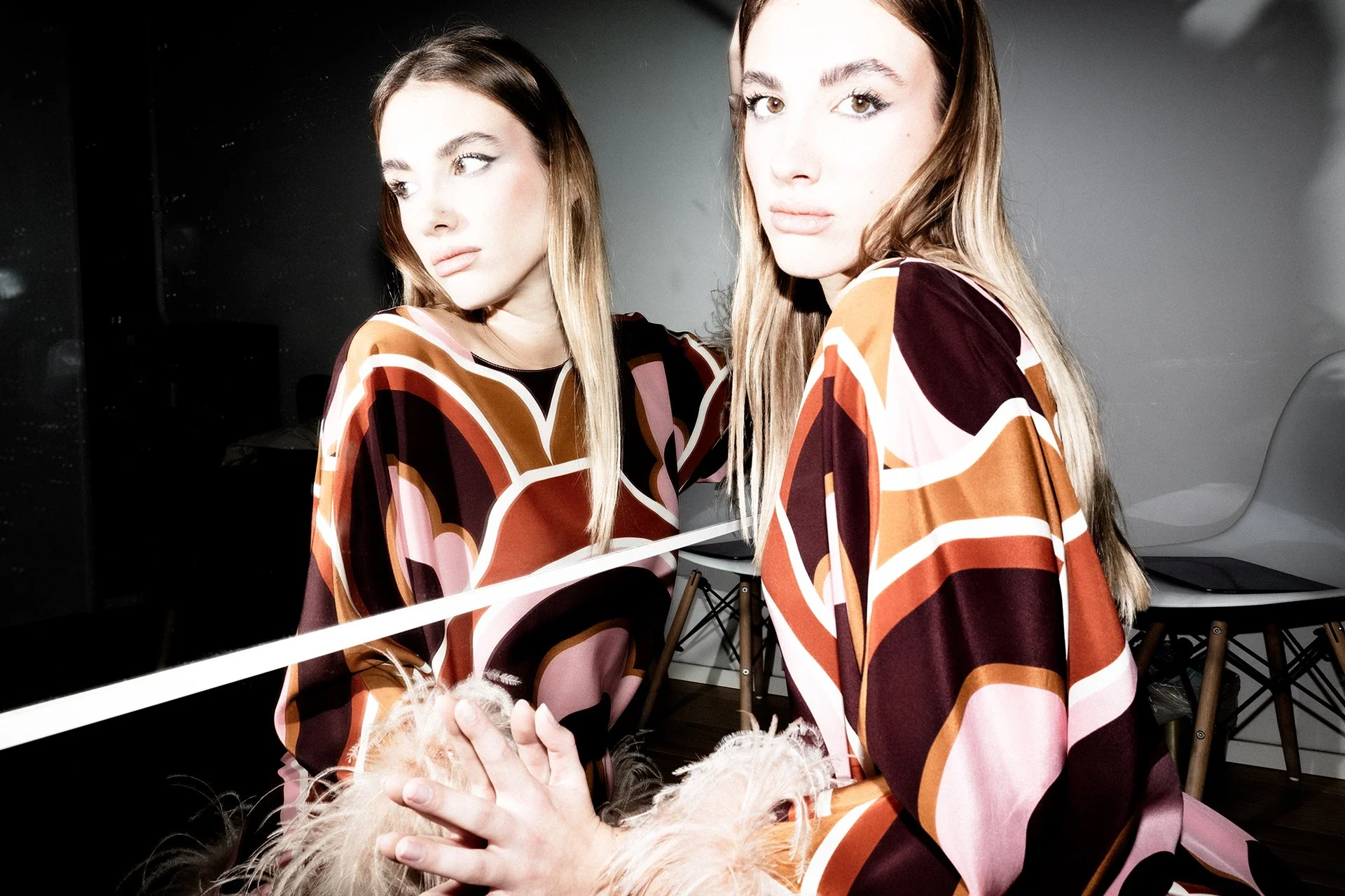 A woman with long blonde hair looking at her reflection in a mirror, wearing a patterned blouse with orange, pink, and brown tones, holding a fluffy pink purse.