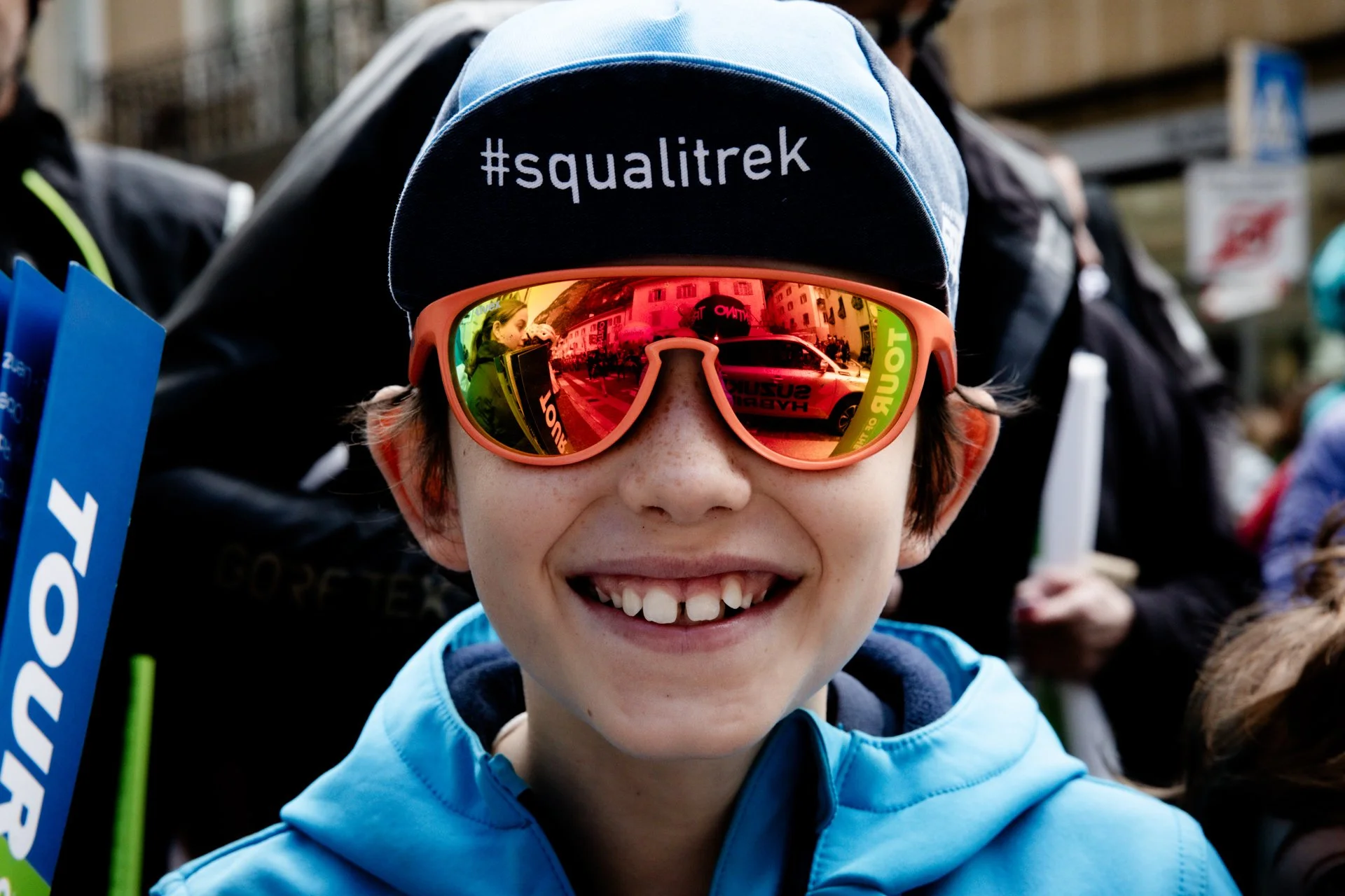 A smiling boy wearing reflective sunglasses, a black and blue cap with '#squalitrek' written on it, and a blue jacket, at an outdoor event.