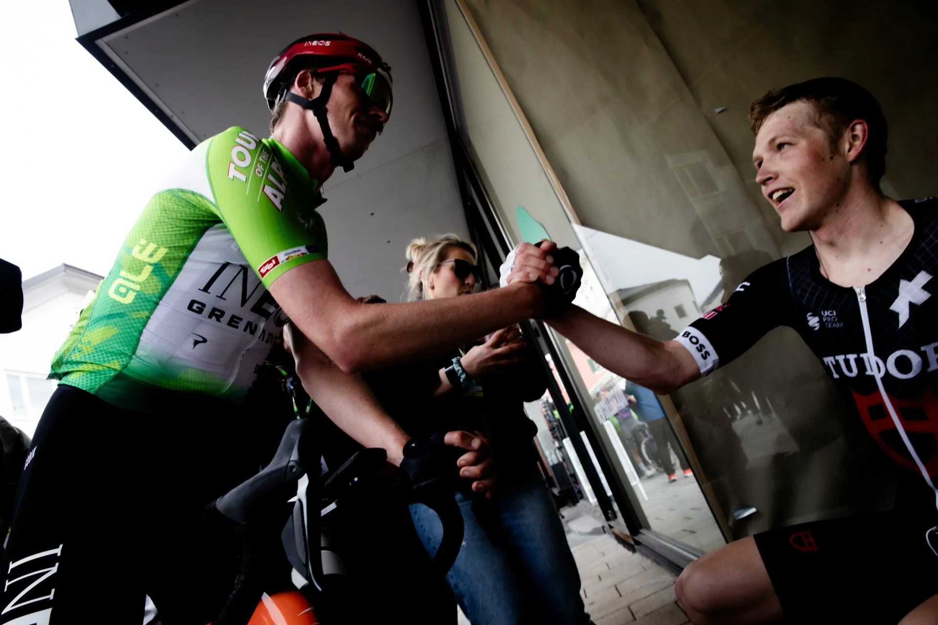 Cyclist in green and white jersey shaking hands with another cyclist in black jersey outside building.