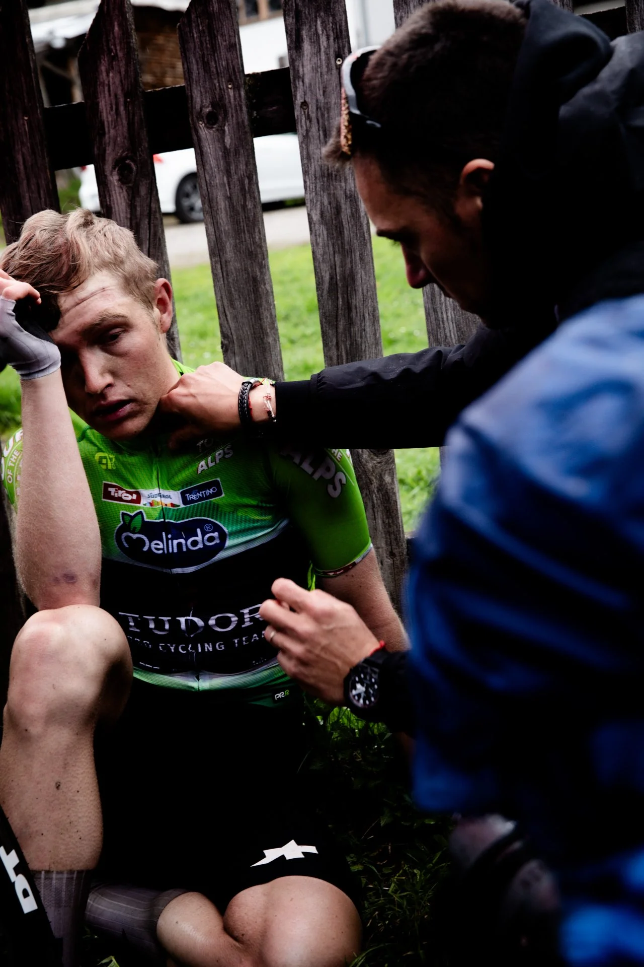 A cyclist is seated against a wooden fence, appearing distressed, as a person tenders to his injury or provides medical assistance.