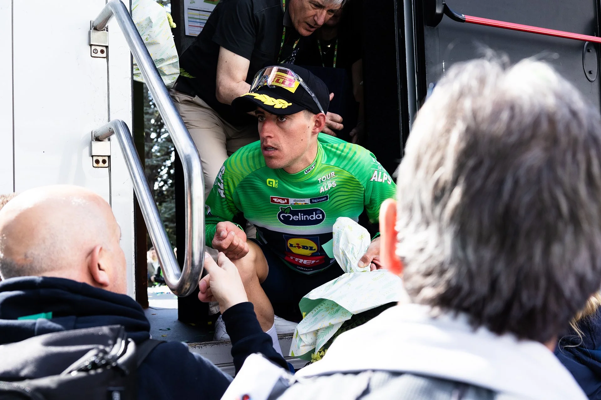 A cyclist in a green jersey with various sponsor logos, wearing a black cap with yellow and gray stripes, sitting at the back of a vehicle, interacting with fans. A man in a black shirt leans inside the vehicle, and several fans with gray or bald hea