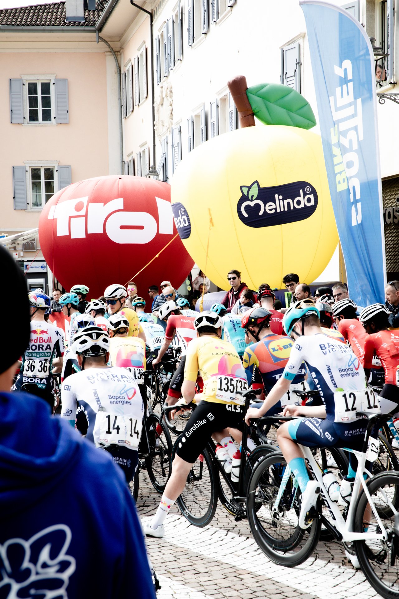 Cyclists at the starting line of a race, with large inflatables and a blue flag with the words 'Bike Store' in the background.
