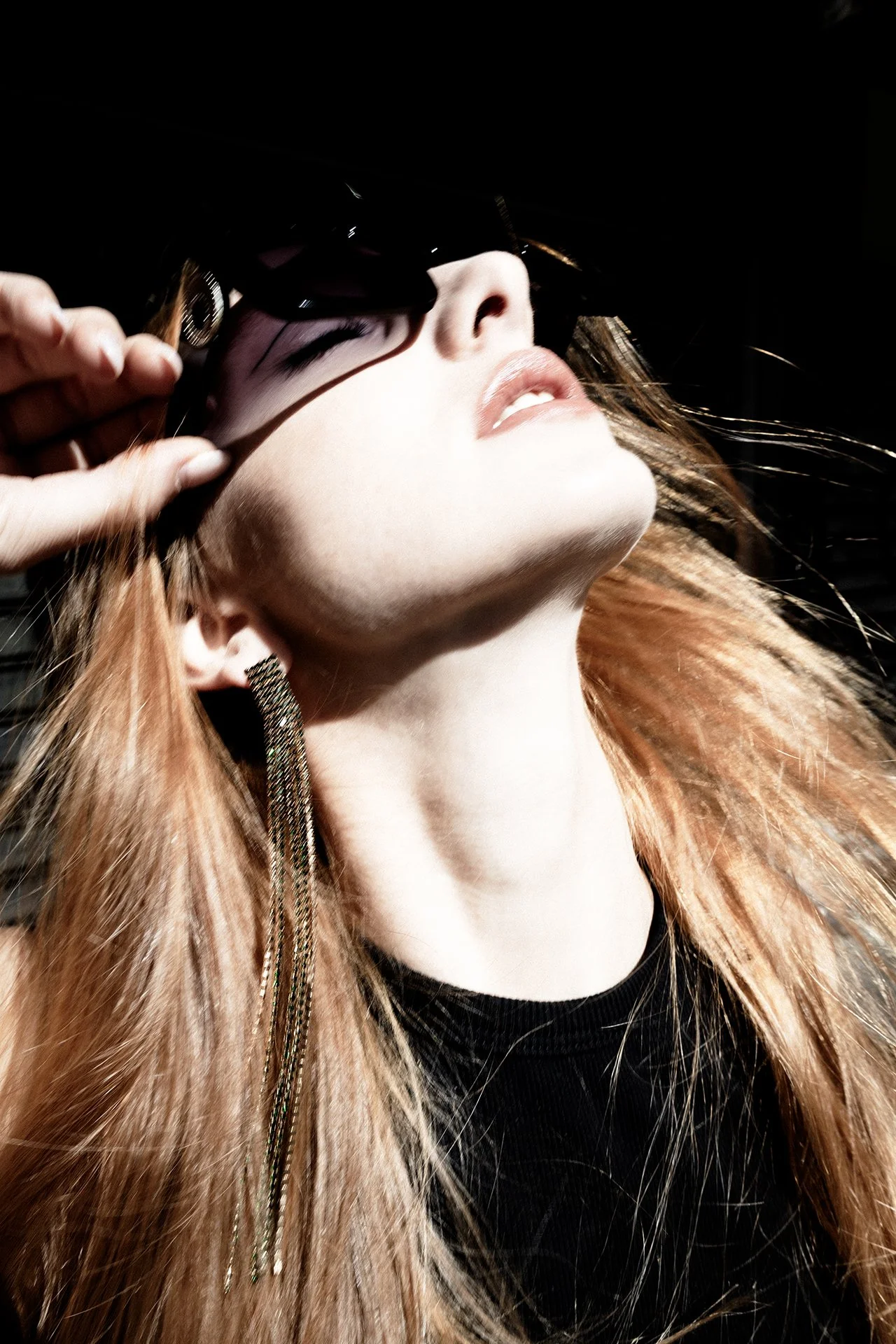 A woman with long red hair, wearing large dangle earrings, raises her head slightly with her eyes closed, as someone adjusts her black sunglasses.