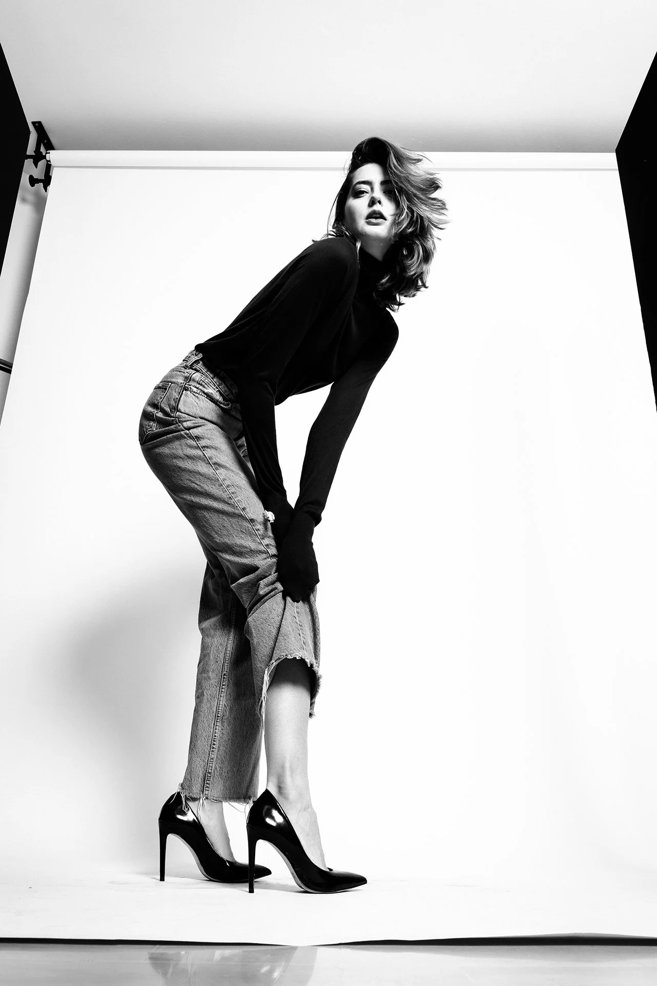 A woman with wavy hair wearing a black top, ripped jeans, and high heels posing in front of a white backdrop in a photo studio.