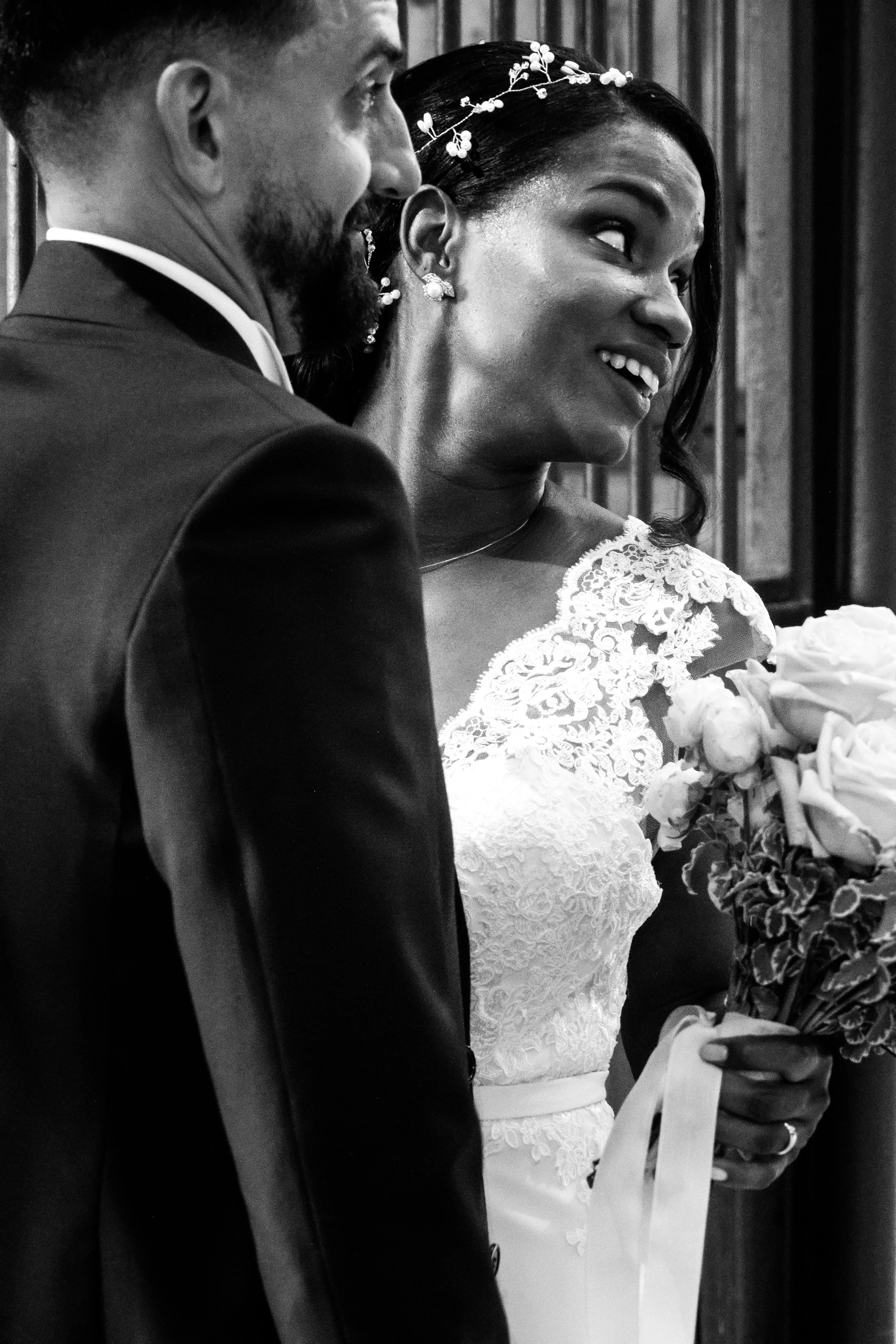A black and white photo of a smiling bride holding a bouquet of flowers, looking at her groom during their wedding ceremony.