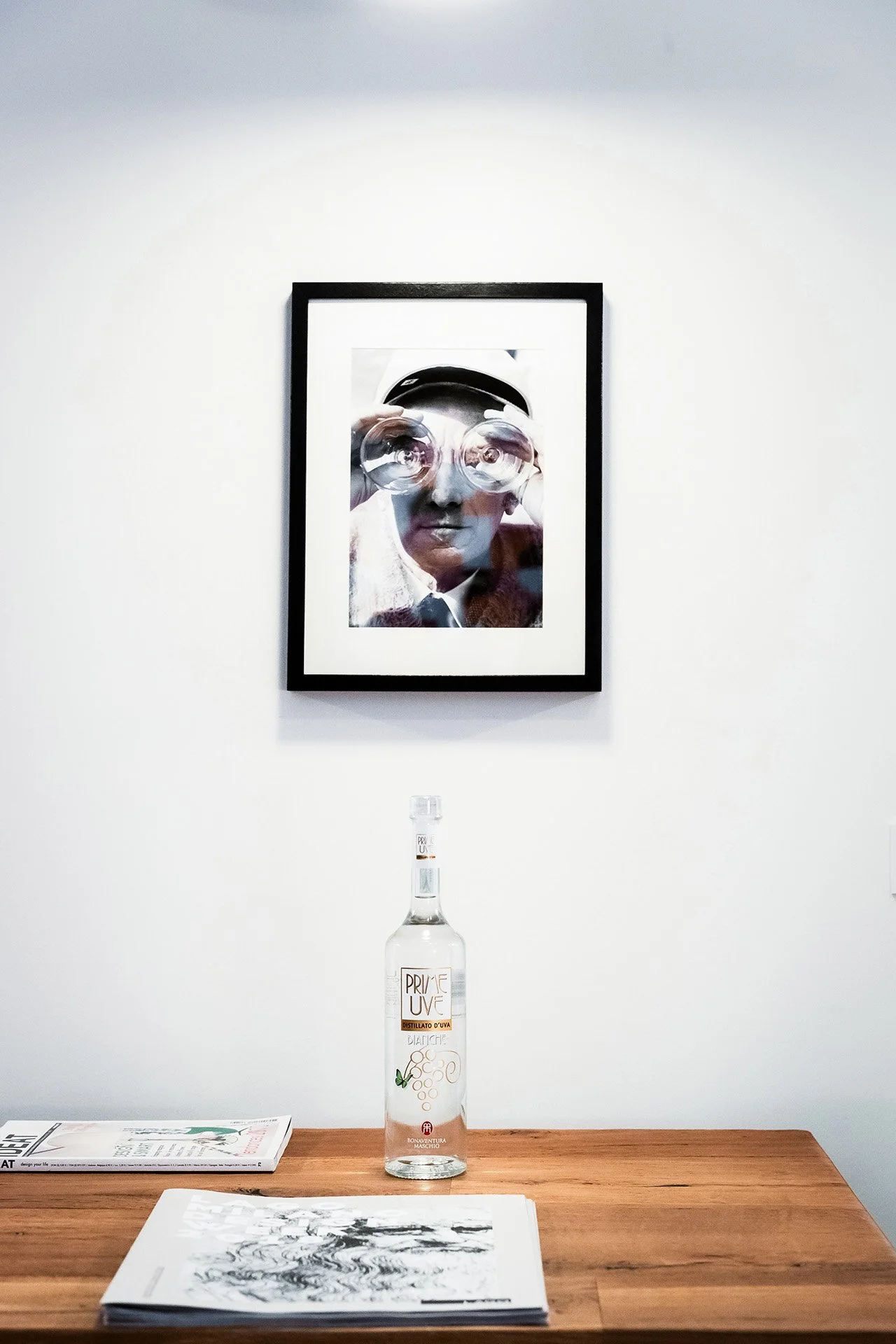 A framed black-and-white photo of a man holding glasses in front of his face, hanging on a white wall. Below, on a wooden table, there is a clear bottle of Prime Uve grape distillate and some papers or magazines.