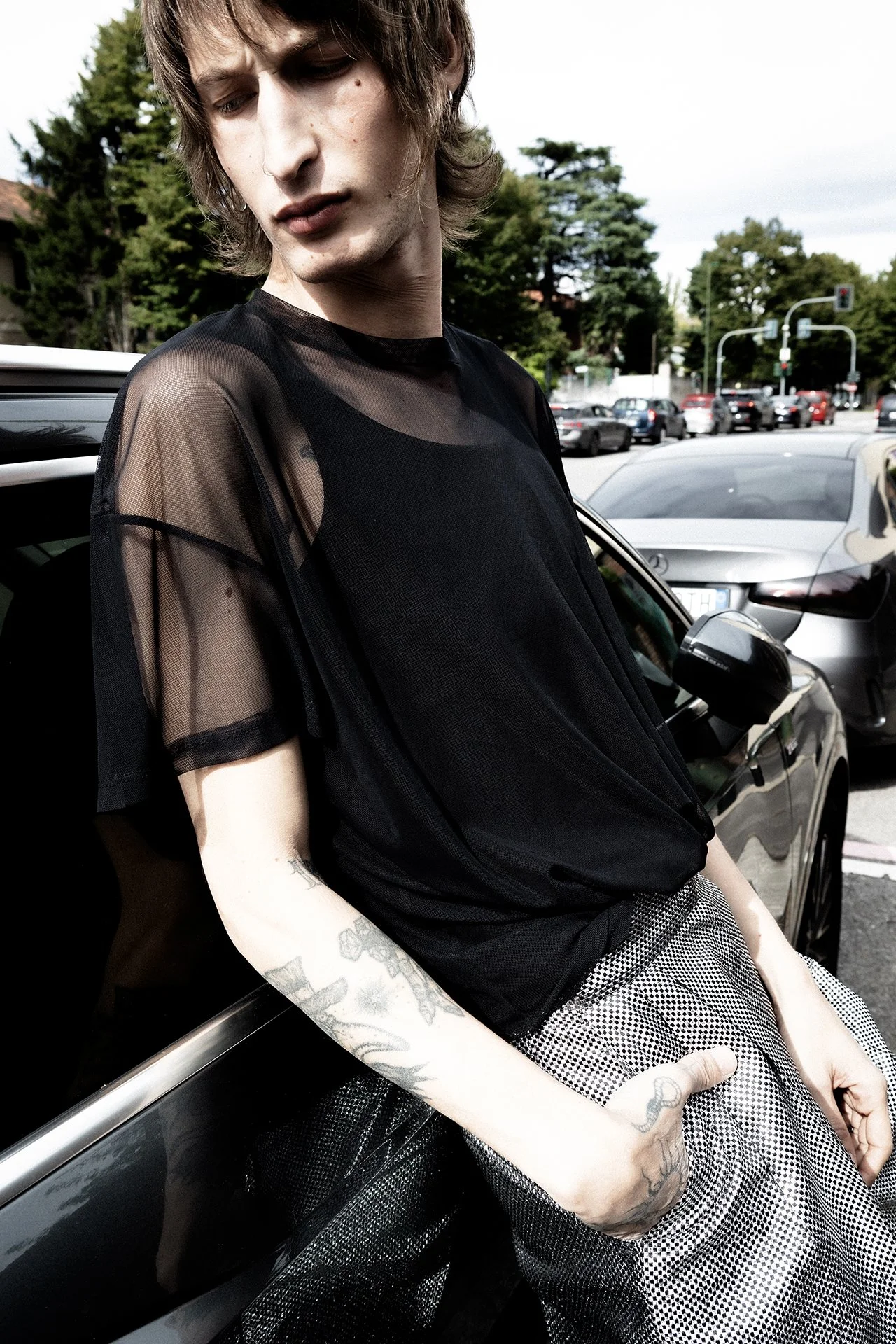 A young man with tattoos on his arms, wearing a black sheer t-shirt and patterned pants, leaning against a black car in a parking lot during daytime.