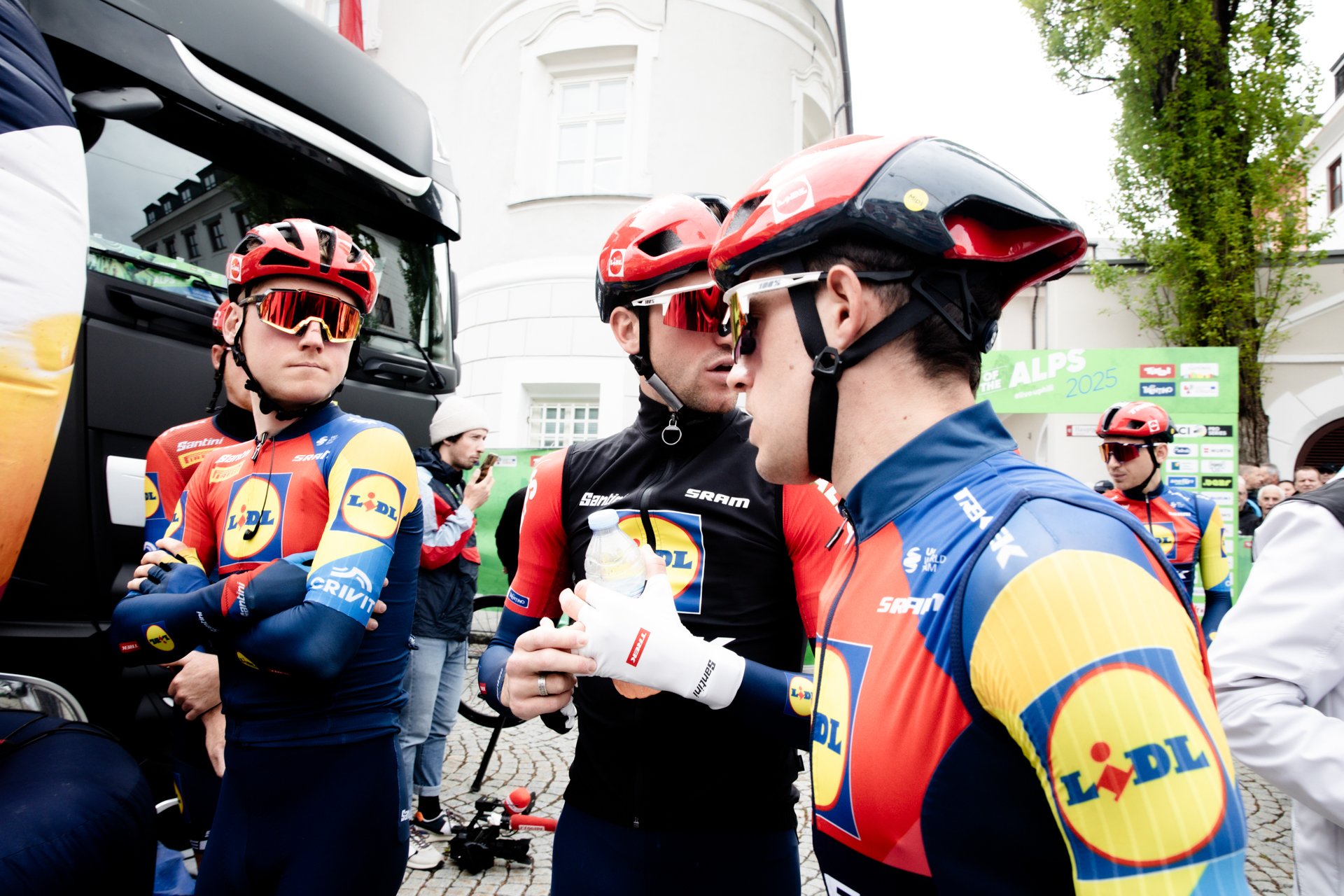 Cyclists in matching Lidl team uniforms, wearing helmets and sunglasses, gathered together during a race event, with some holding water bottles and others engaging in conversation.